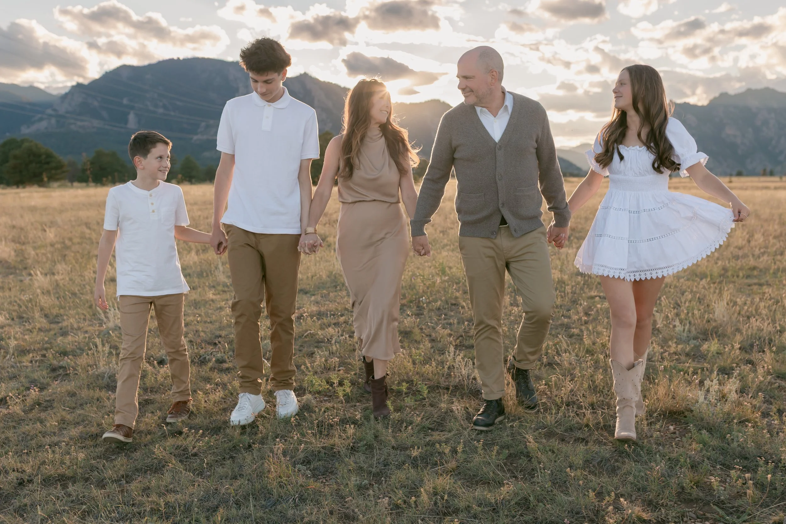 A family of six walking hand in hand outdoors during sunset in a grassy field, with mountains in the background.