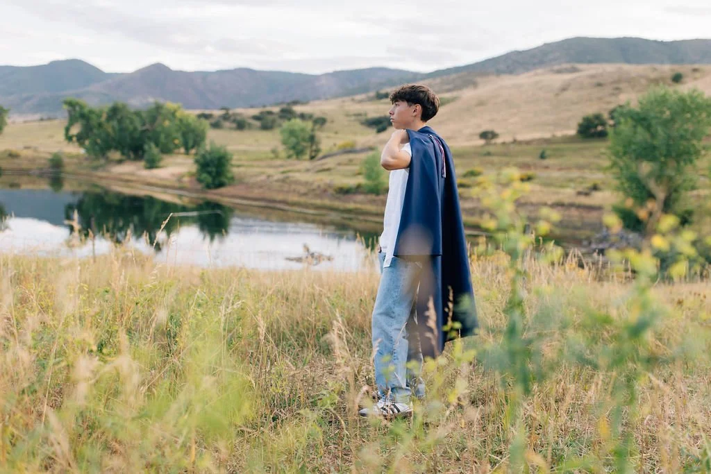 Young man holding his jacket over his shoulder, standing in a field near a lake with mountains in the background.