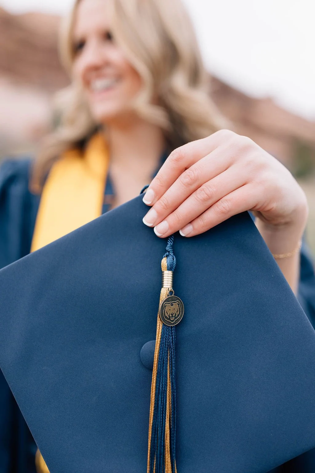 Close-up of a woman holding a graduation cap with a tiger emblem tassel in front of her, with a blurred smiling woman in a graduation gown in the background.