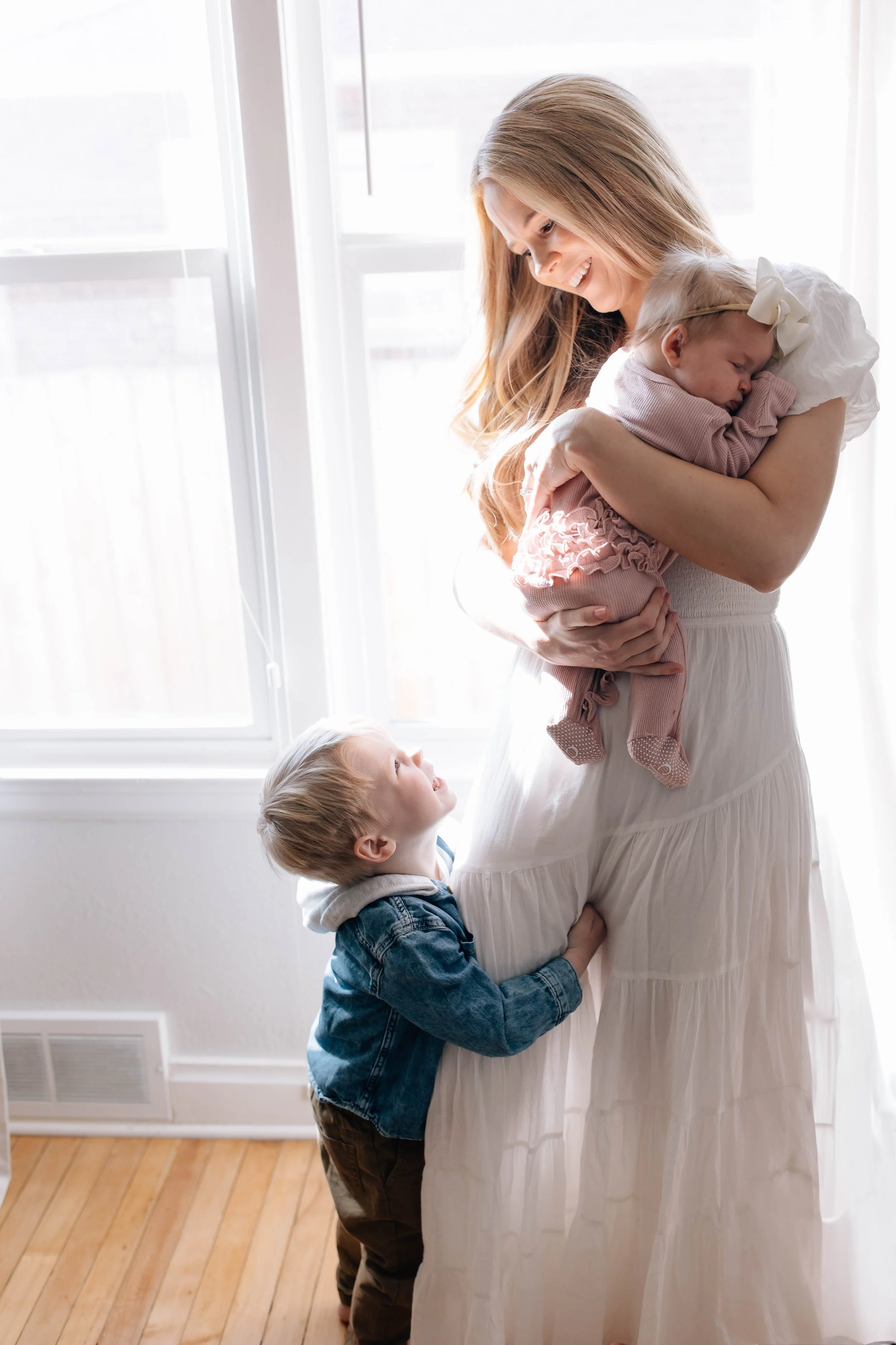 A woman holding a sleeping baby girl while an older boy hugs her leg, looking up lovingly at her near a window.