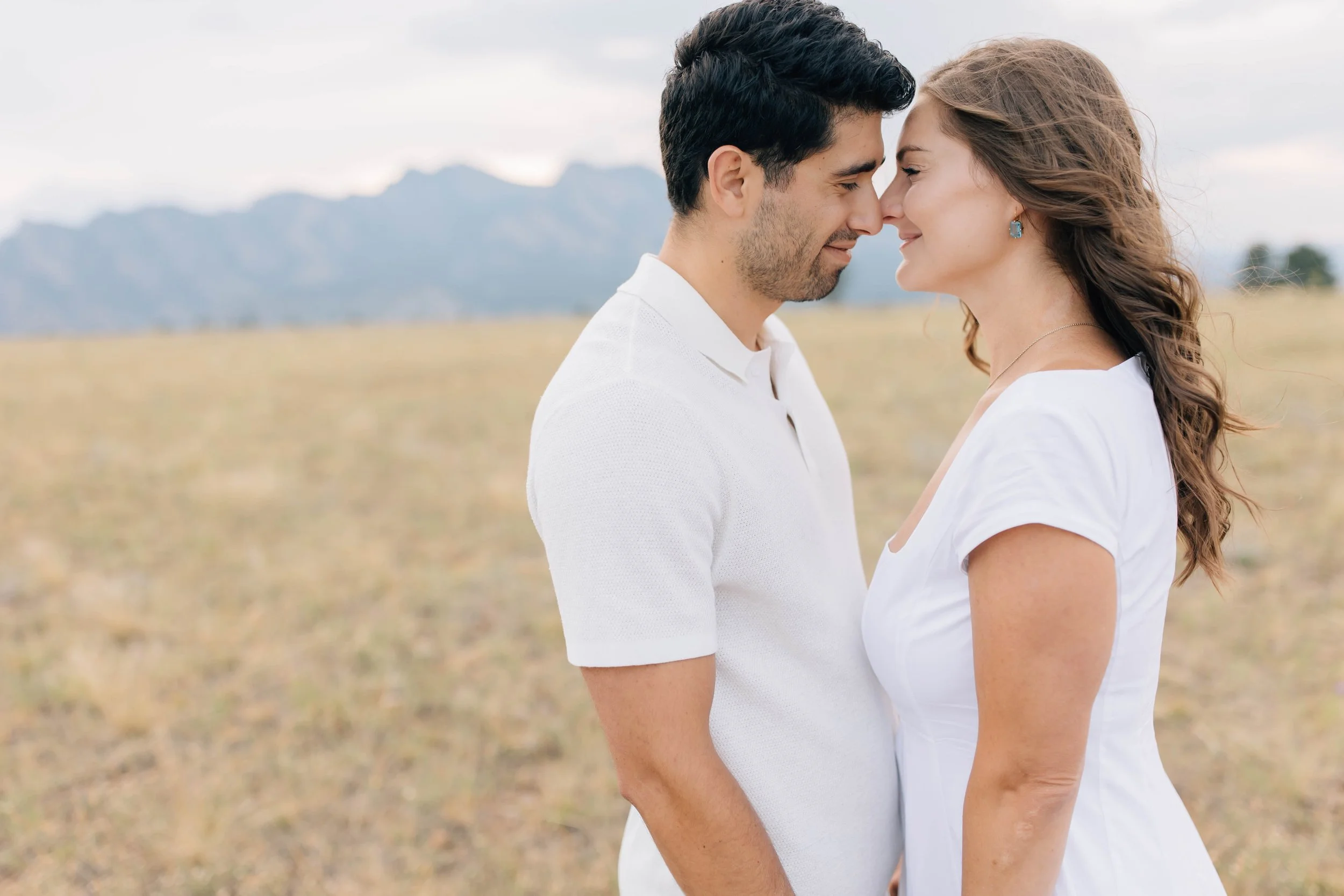 A man and woman standing close together outdoors with their foreheads touching, smiling, in a field with mountains in the background.
