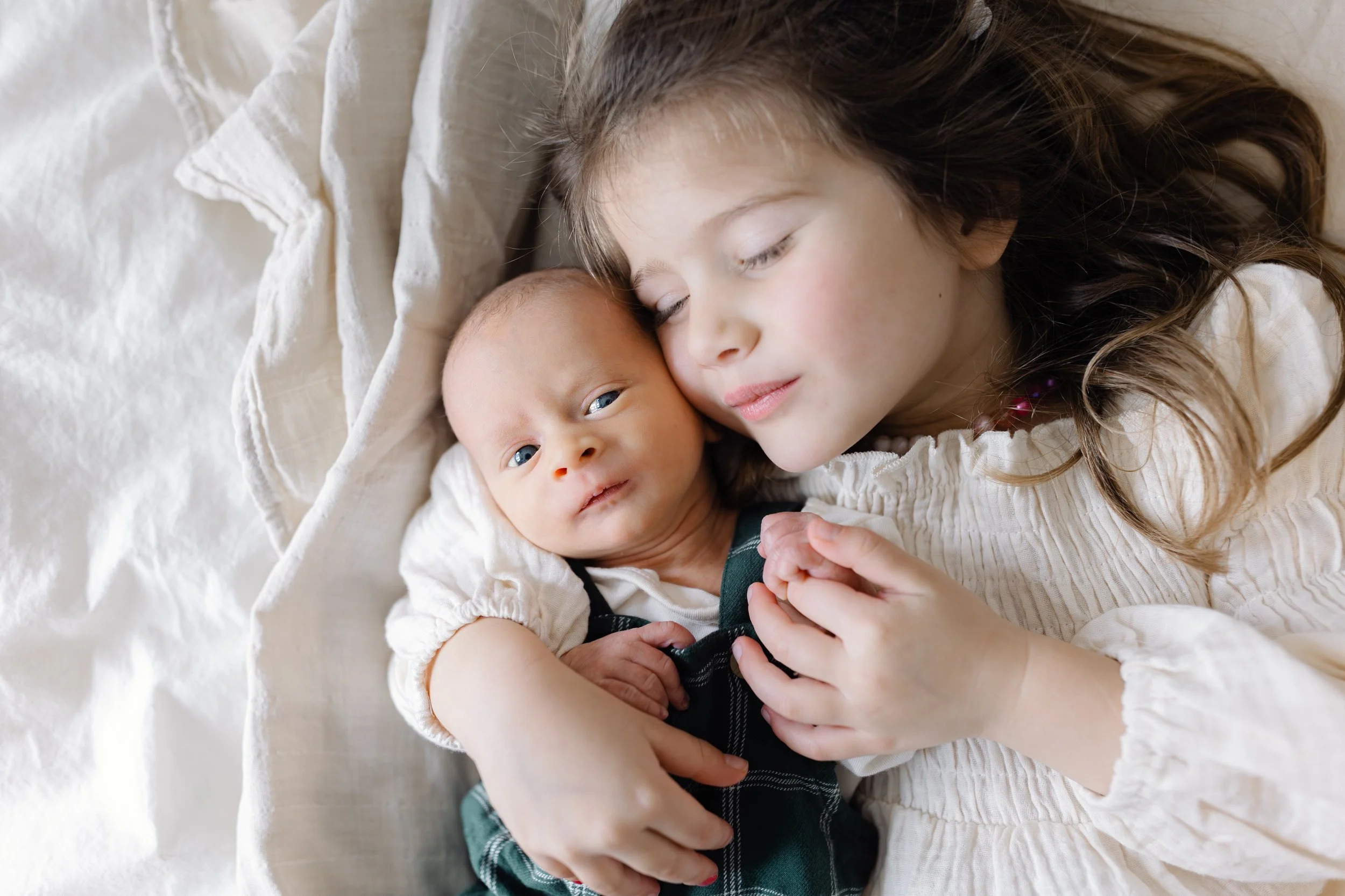 A young girl with long brown hair and light skin resting her head on a baby with light skin, lying on a bed. The girl has closed eyes and the baby is looking at the camera with a curious expression.