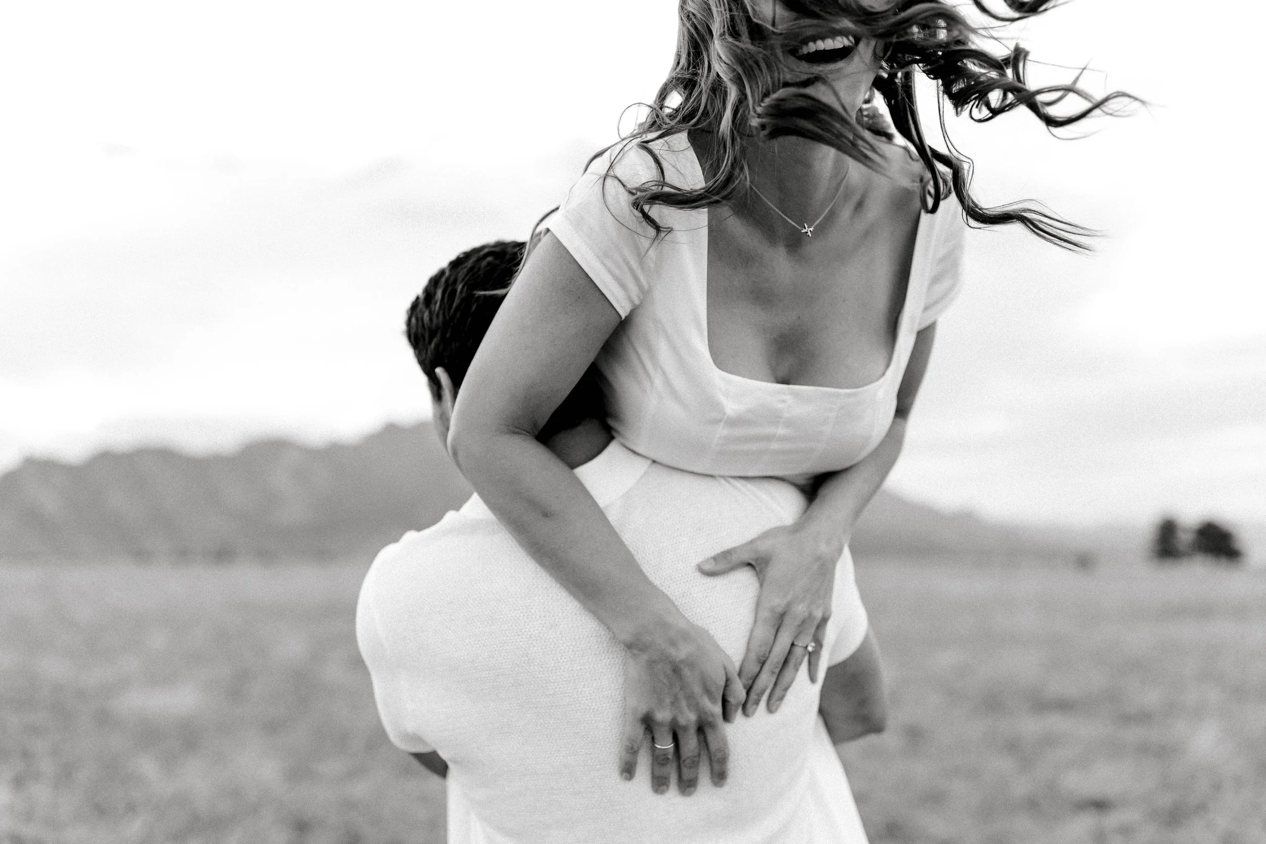 A woman with long hair, wearing a necklace and a white top, is being carried on the shoulders of a man in an outdoor landscape with mountains in the background. The image is in black and white.