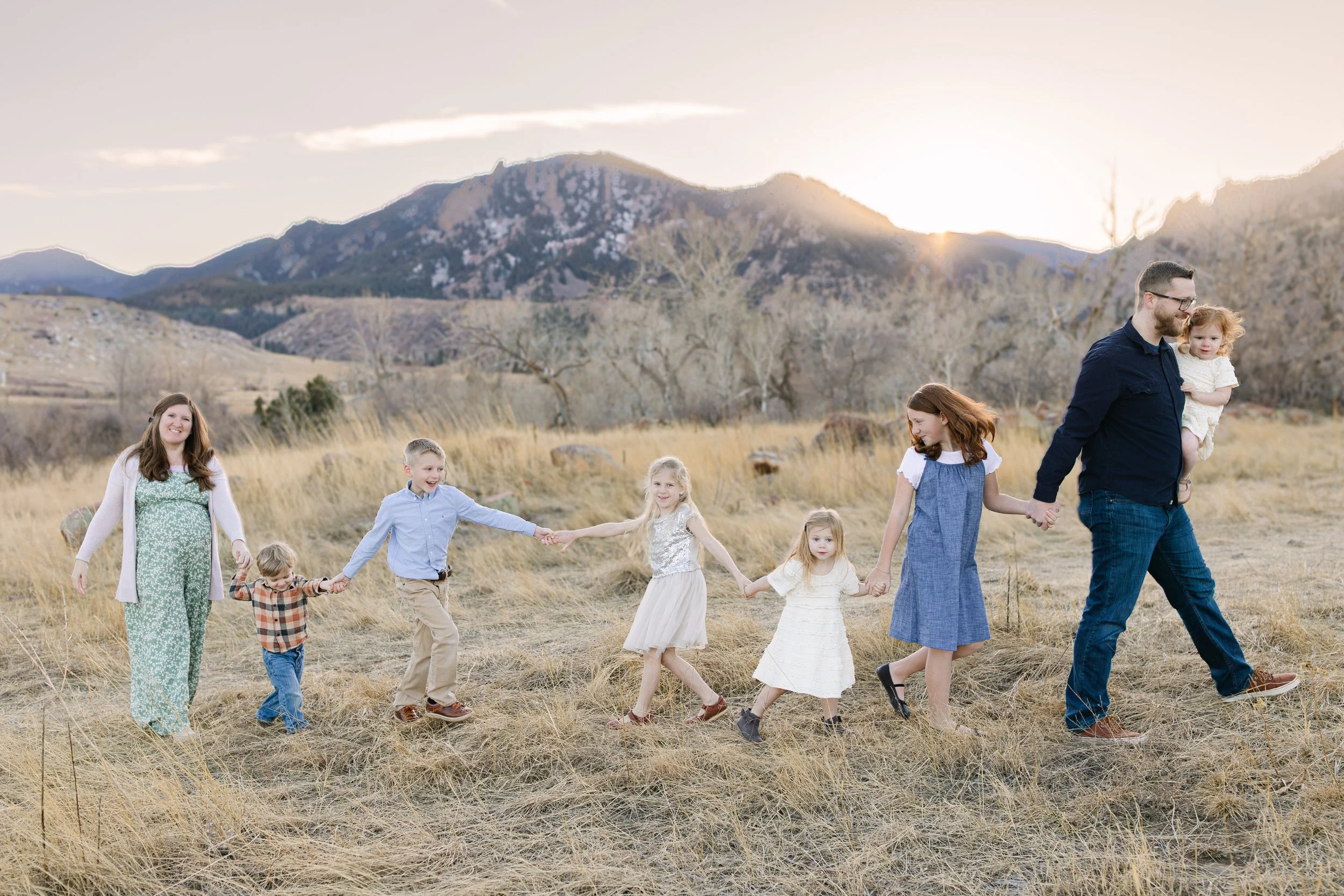 Family of nine holding hands and walking across a grassy field with mountains in the background at sunset.