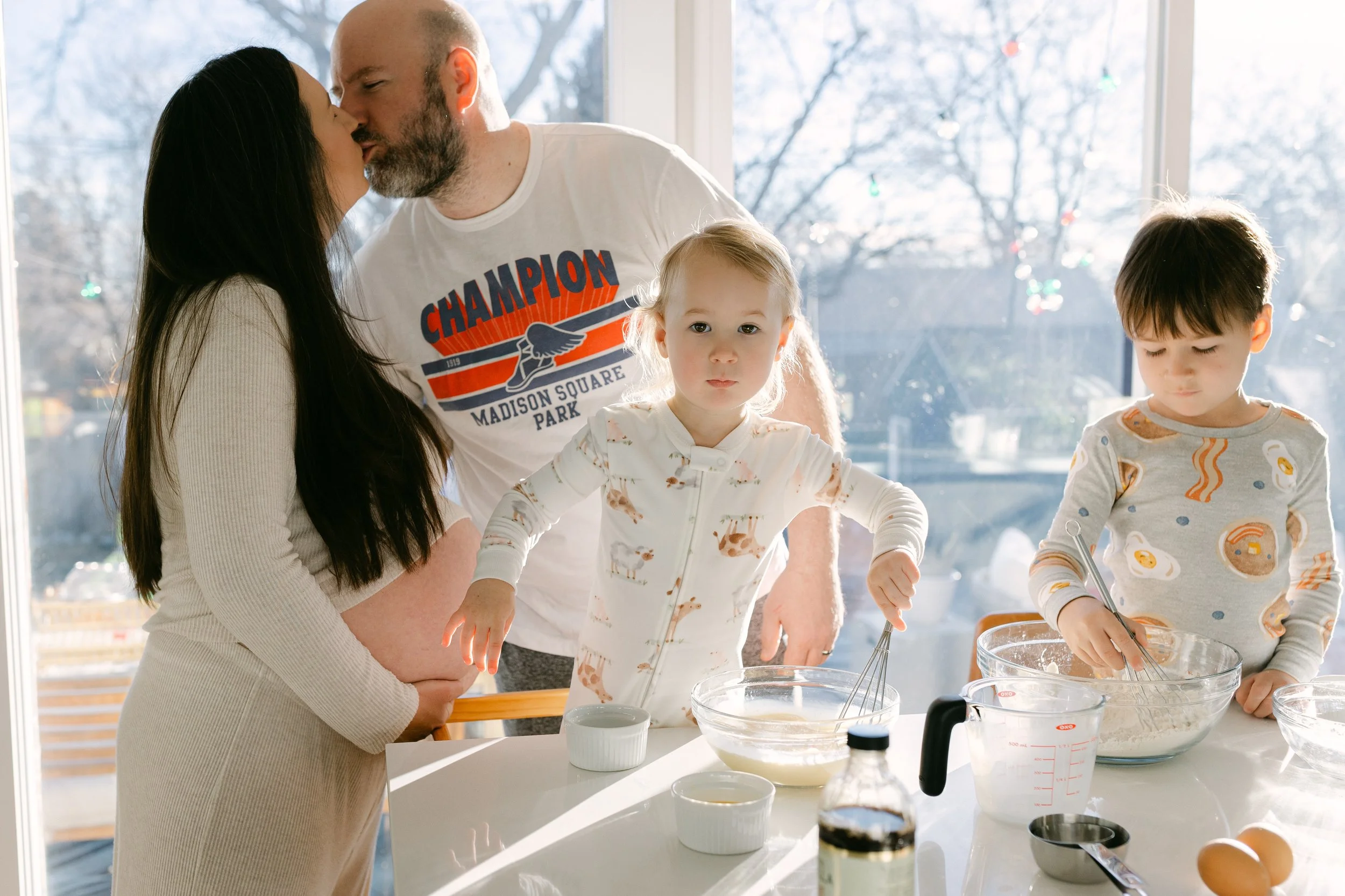 A family in a kitchen, with a woman pregnant, a man and two young children, engaging in baking. The woman and man are kissing near the window, the children are mixing ingredients in bowls on the table, which also has eggs, measuring cups, and kitchen