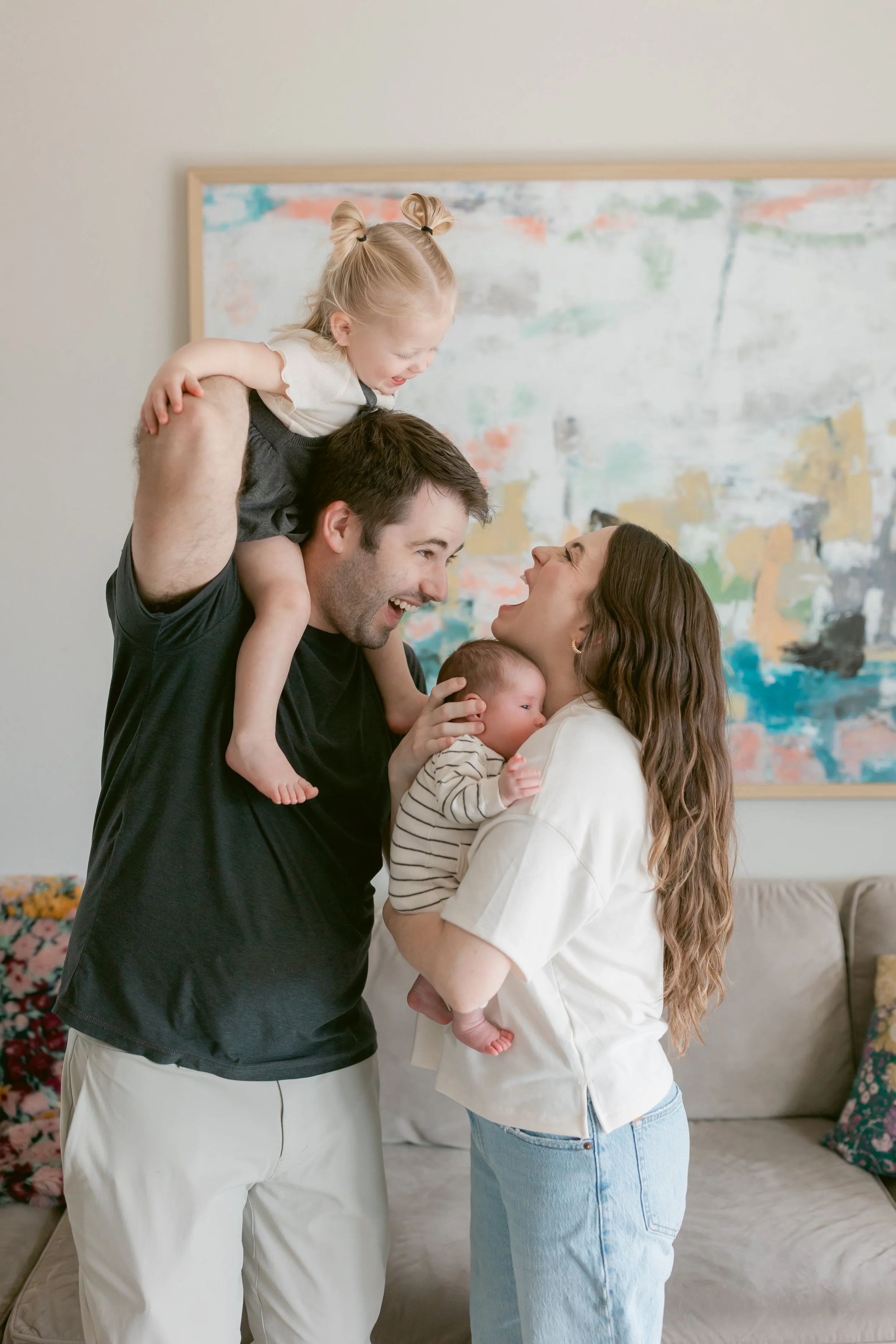 A family of four, including a father holding a young girl on his shoulders, a mother holding a baby, are smiling and interacting joyfully indoors in front of an abstract painting.