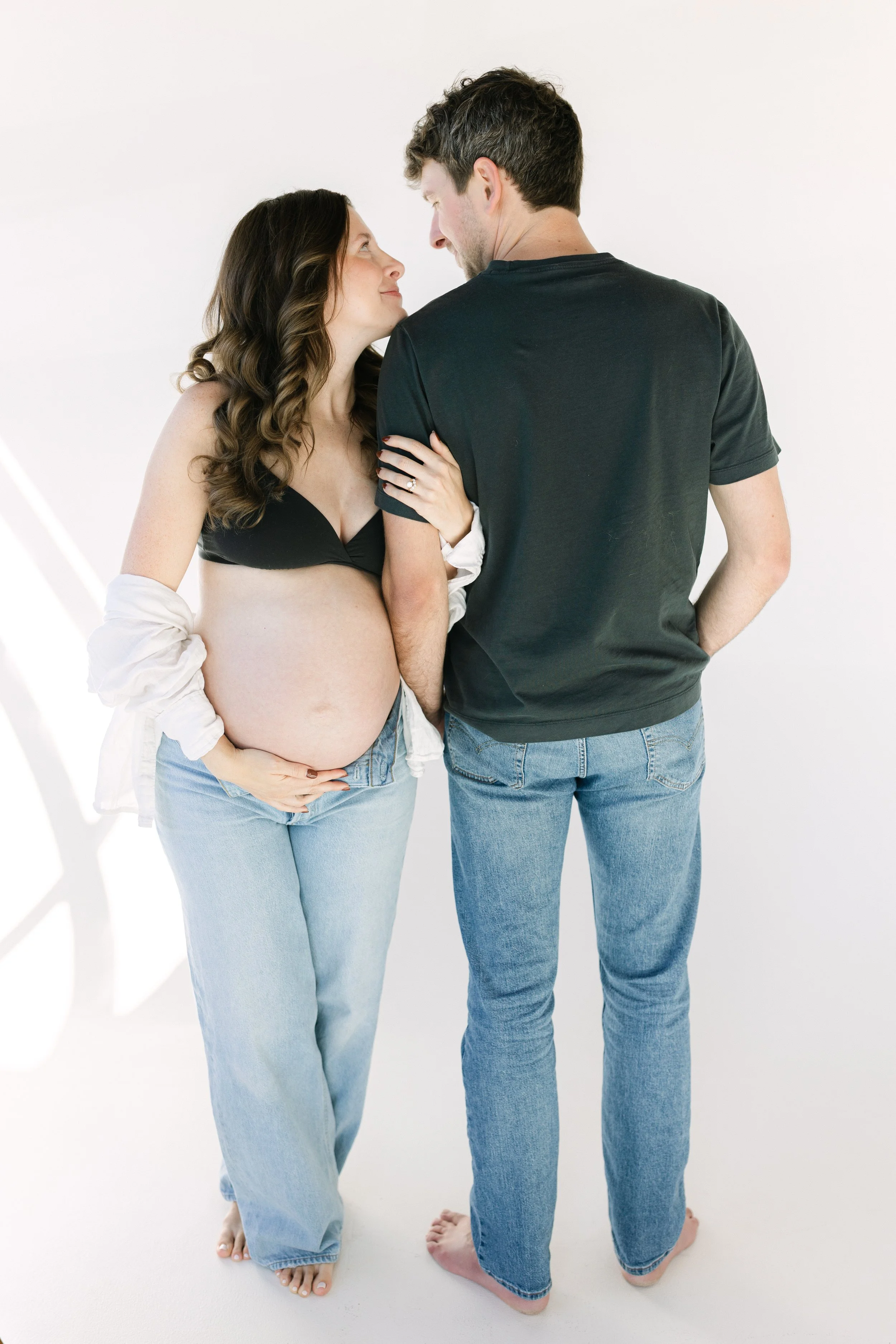 A pregnant woman and a man standing close together, looking into each other's eyes, in a bright, minimalist setting.