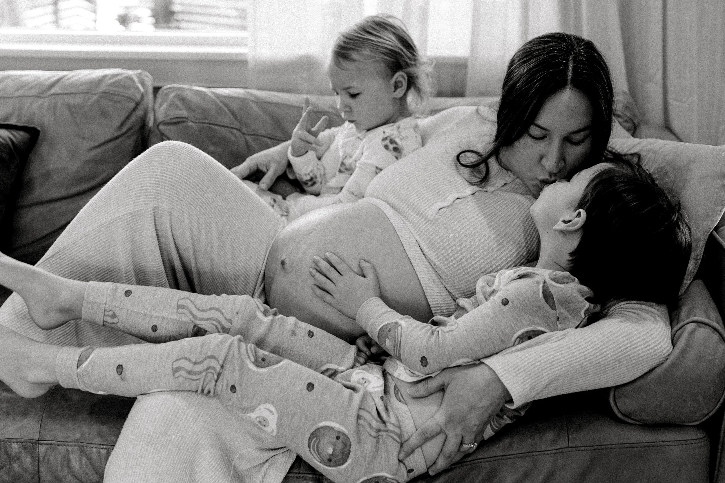 A pregnant woman lying on a couch with two children, one child sitting on her lap and the other one leaning against her, touching her belly, in a cozy living room.