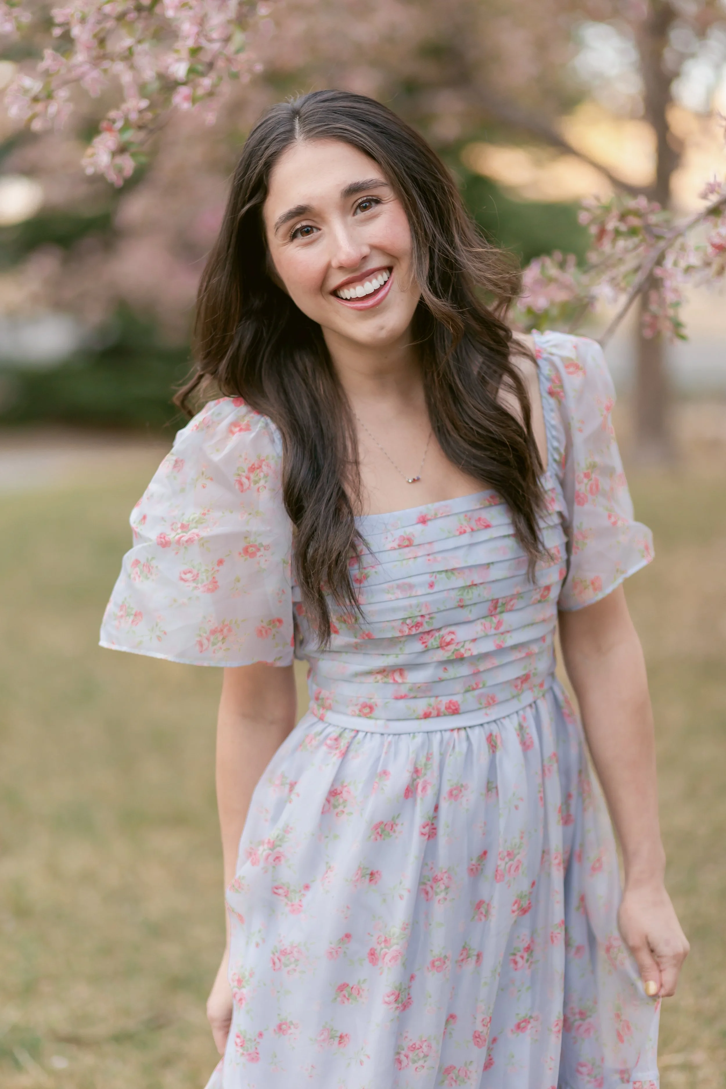 A young woman in a floral dress standing outdoors near blooming pink cherry blossom trees, smiling at the camera.