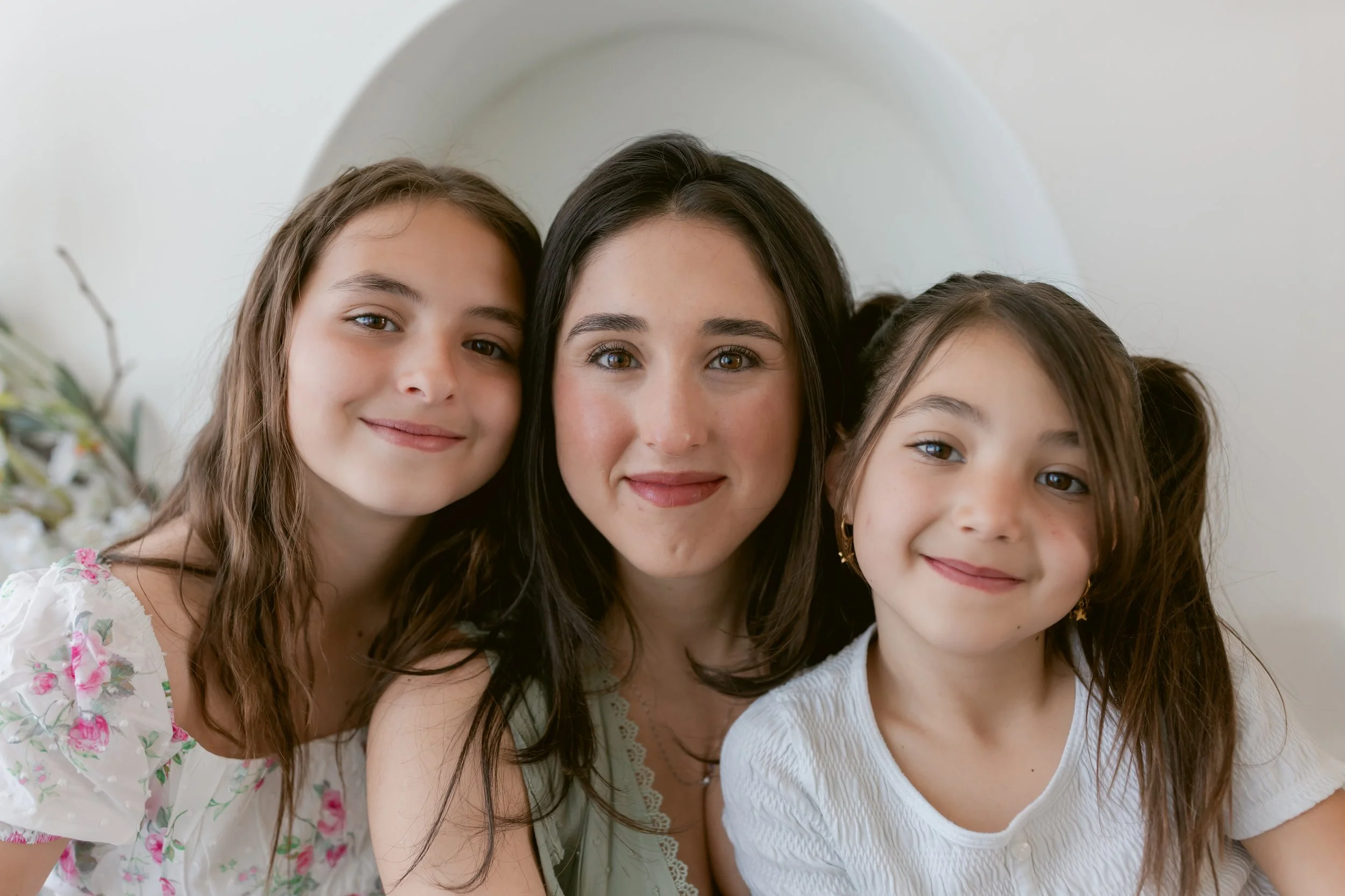 A woman with two young girls, all smiling and close together, in a bright indoor setting.