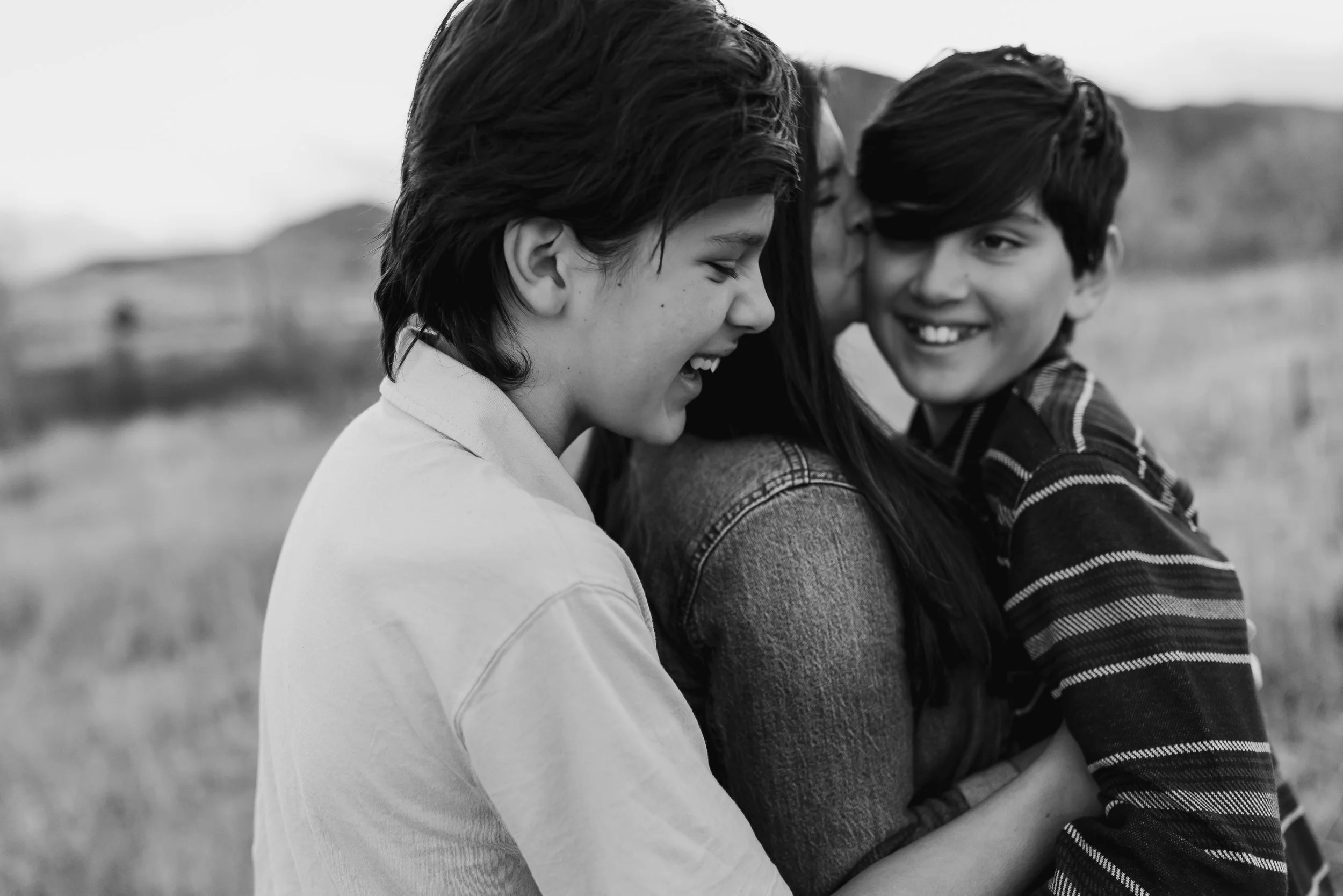Three people, a woman with two children, are outdoors and sharing a joyful moment, hugging and smiling