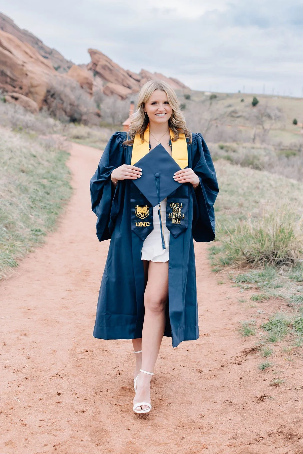 A young woman in a graduation gown and cap standing on a dirt path outdoors, smiling and holding her cap, with rocky hills in the background.