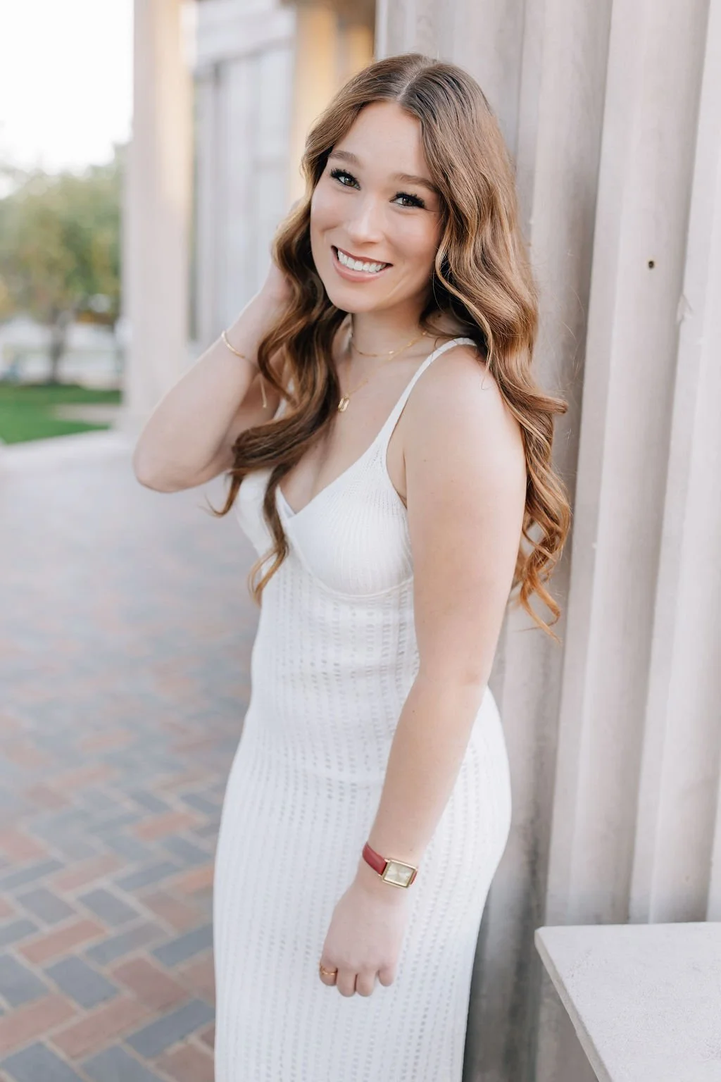 A young woman with long, wavy auburn hair smiling, wearing a white sleeveless dress, standing outdoors beside a white wooden structure, with trees and a brick pathway in the background.