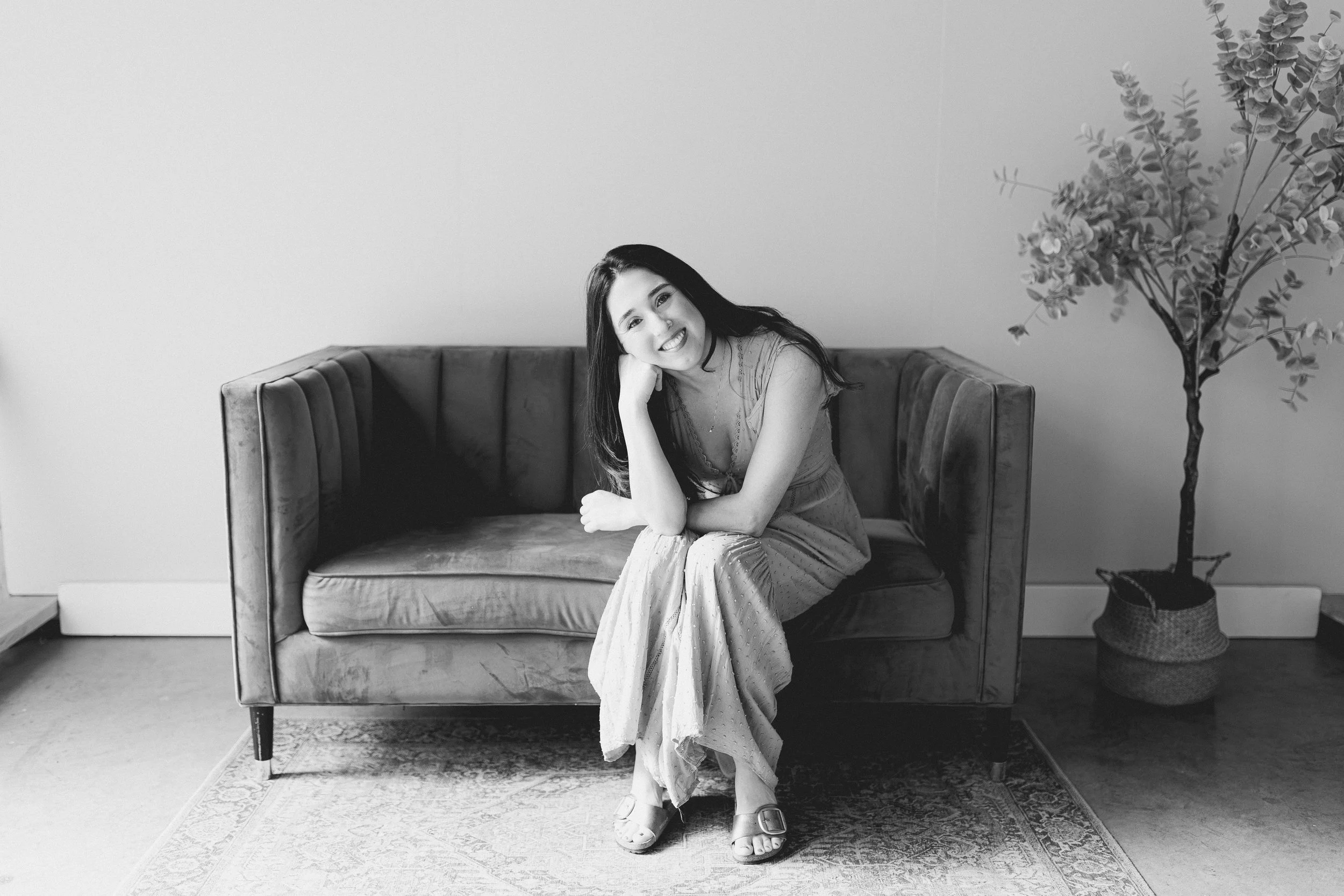 A woman sitting on a velvet sofa, smiling with her head resting on her hand, next to a potted plant in a room with a patterned rug.