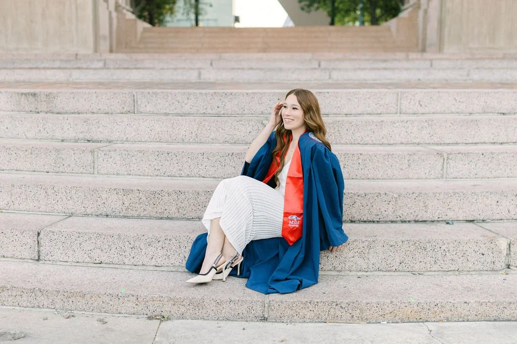 A young woman in a graduation gown and cap sitting on outdoor steps, smiling and posing.