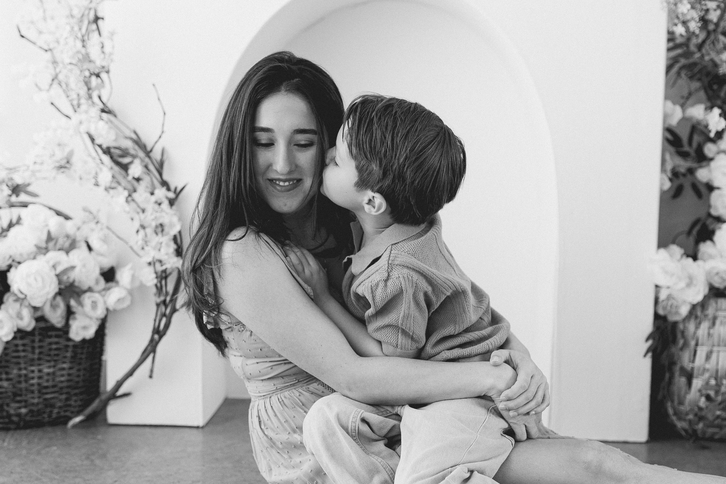 A woman and a young boy share a tender moment, with the boy kissing the woman's cheek, surrounded by floral arrangements.