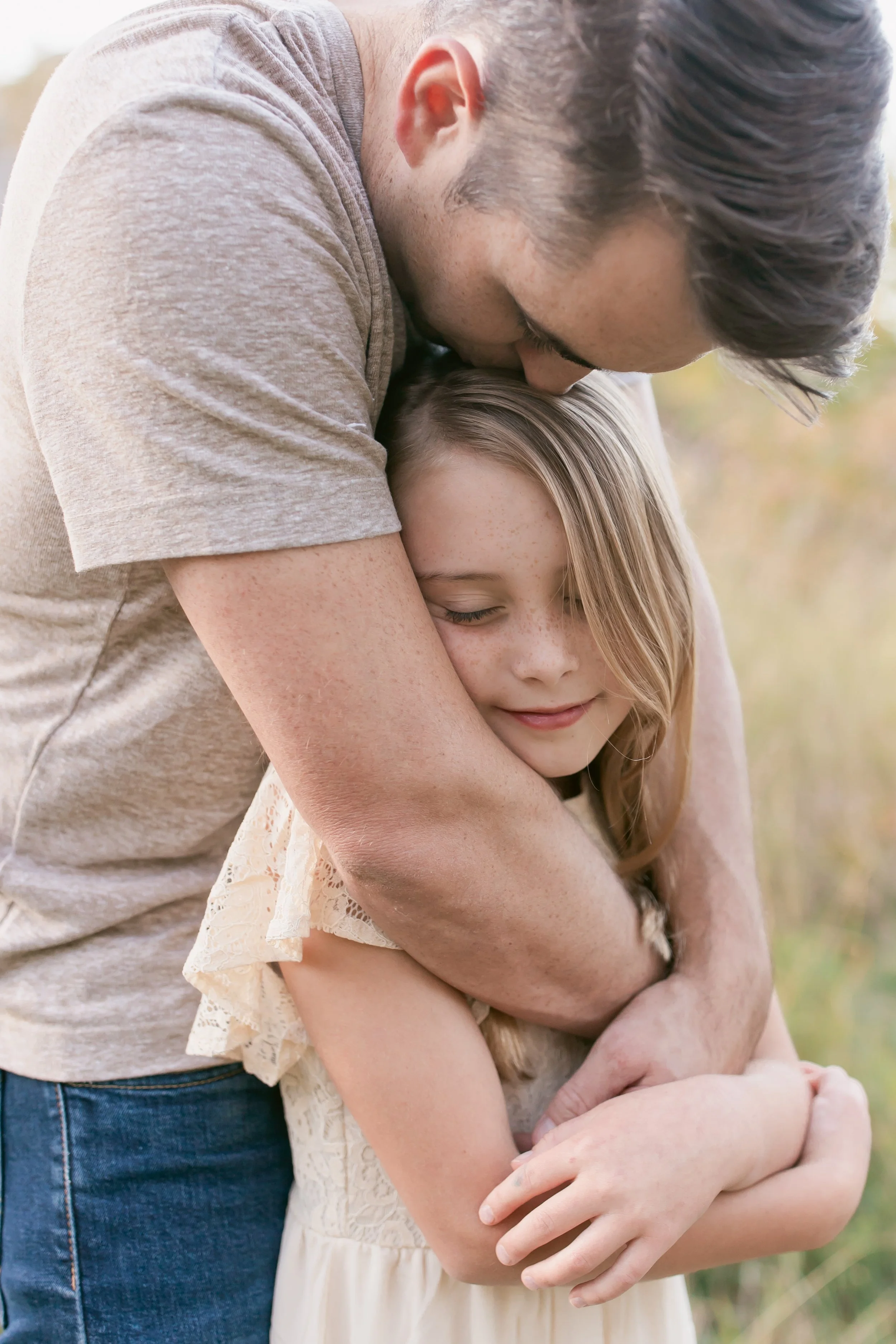 A man embraces a young girl outdoors, his head resting on hers, both with closed eyes and content expressions.
