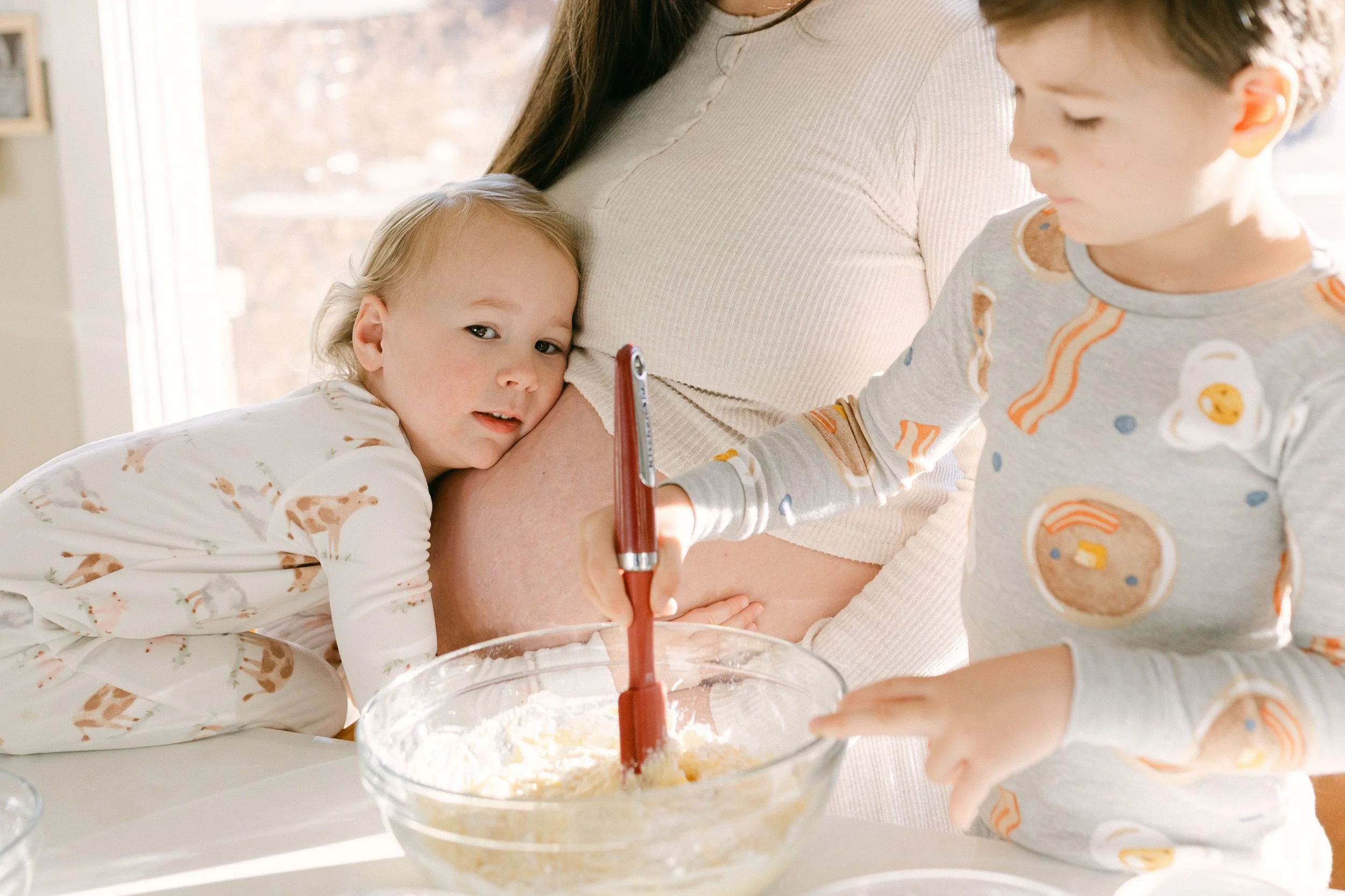 A young girl with blonde hair and pajamas with giraffes hugging her pregnant mother's belly, while a young boy in pajamas with food and drink images stirs a bowl of batter, in a bright kitchen.