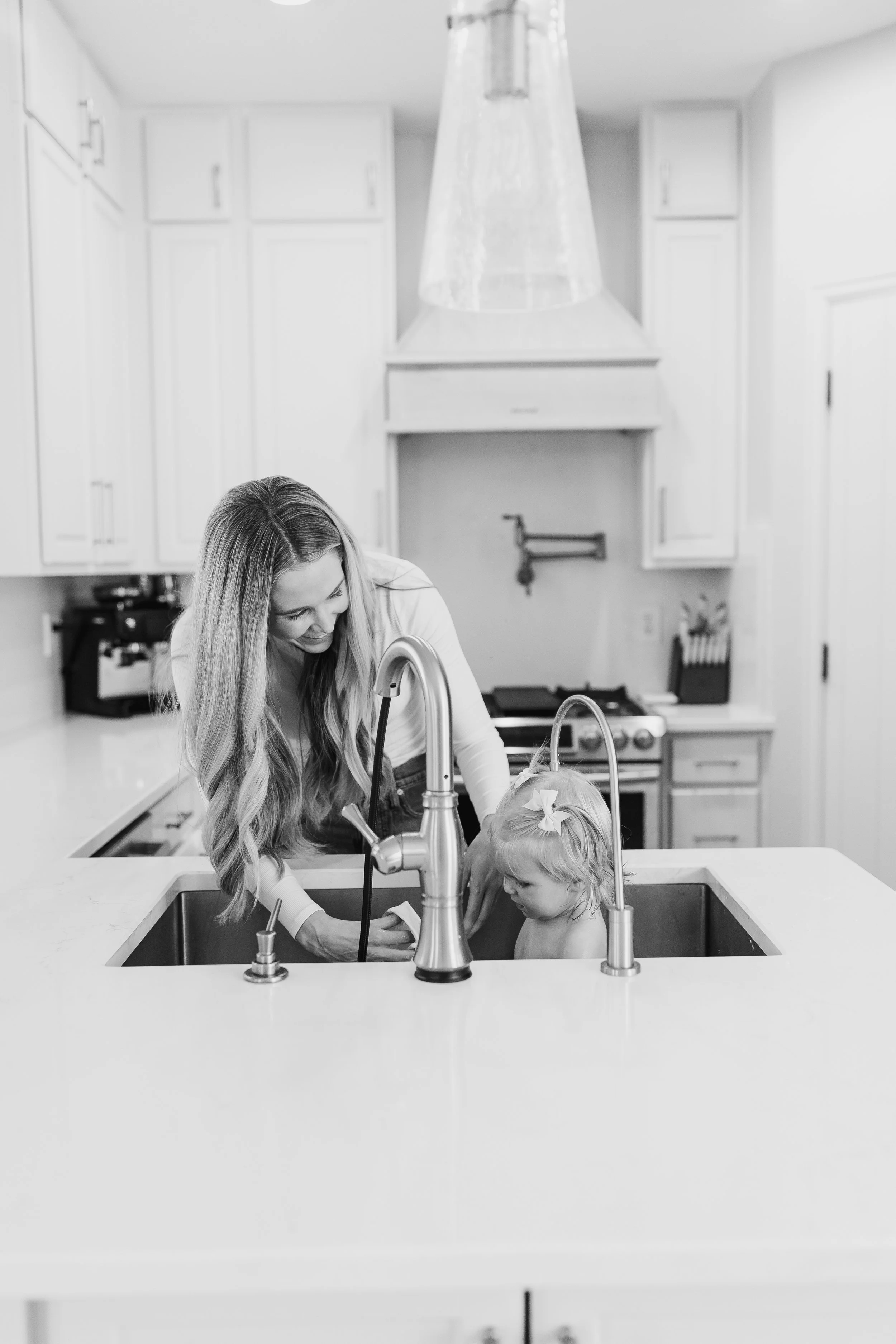 A woman and a young girl standing in a kitchen sink, looking at each other and smiling. The girl has a bow in her hair, and the woman is washing her hands.