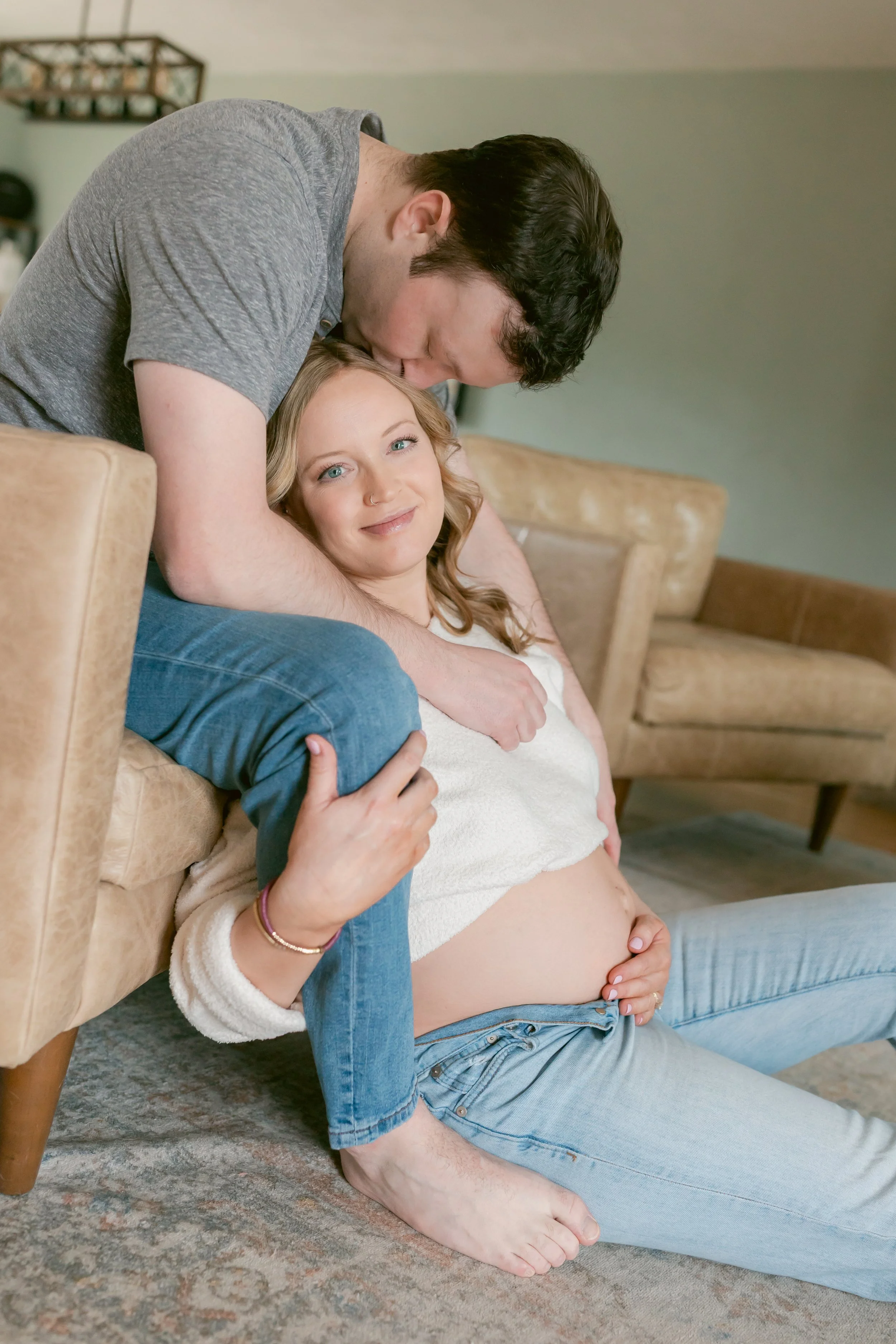 An in-home lifestyle maternity photo of an expecting couple sharing a quiet, intimate moment together. The mother sits comfortably while her partner wraps his arms around her and kisses her head, capturing the warmth, connection, and everyday beauty 