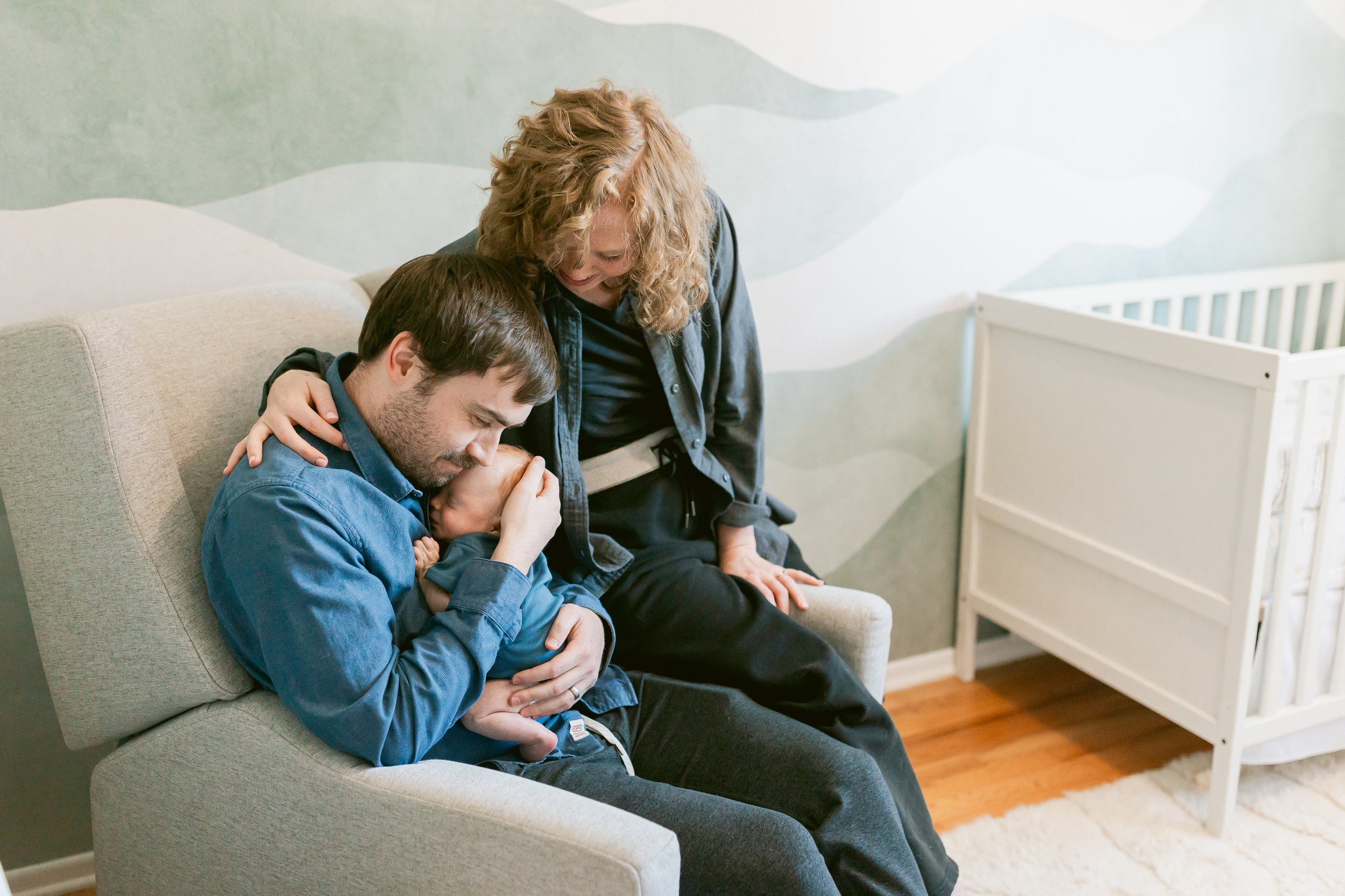 A man sits on a beige couch holding a newborn baby, with a woman standing beside him, all smiling and sharing a tender moment in a nursery.