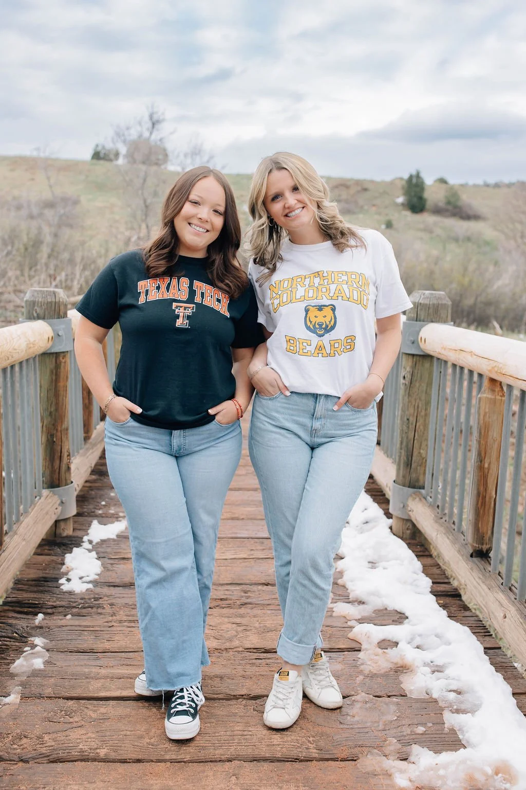 Two young women standing on a wooden bridge with snow on the ground, smiling, wearing university shirts: one University of Texas and the other Northern Colorado Bears, outdoors in a hilly area.