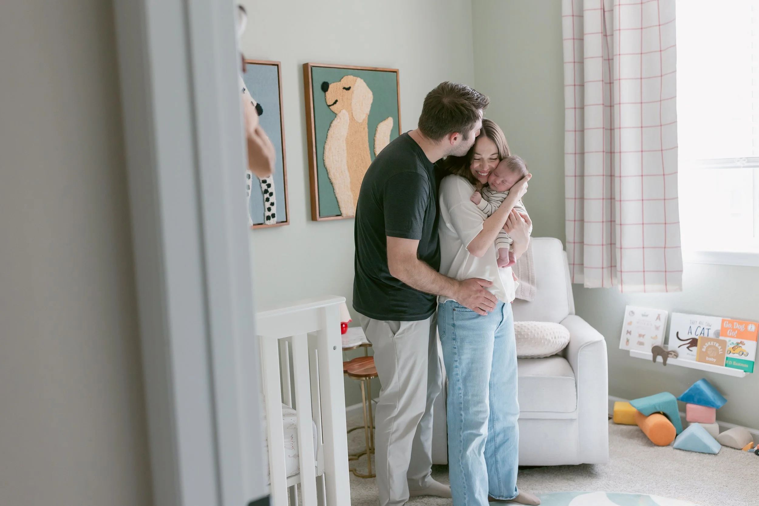 A family of three in a nursery room, with a man, woman, and baby, sharing a joyful moment. The woman is holding the sleeping baby, and the man is leaning in to kiss her.
