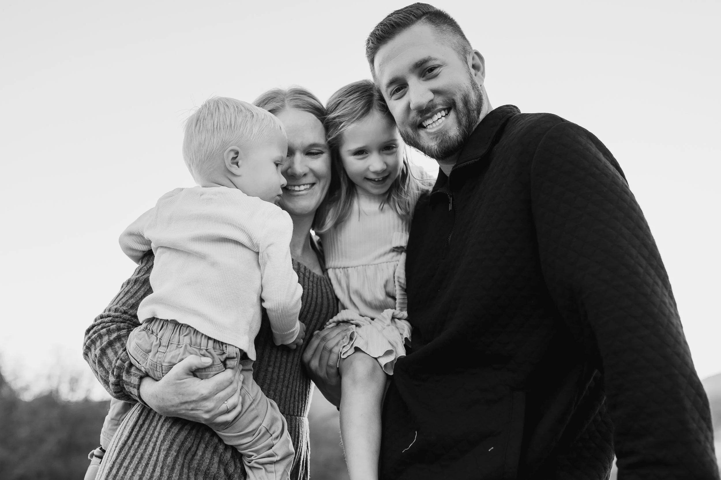 Black and white photo of a smiling family of four outdoors, with a man on the right and woman in the center holding two small kids, a boy and a girl.
