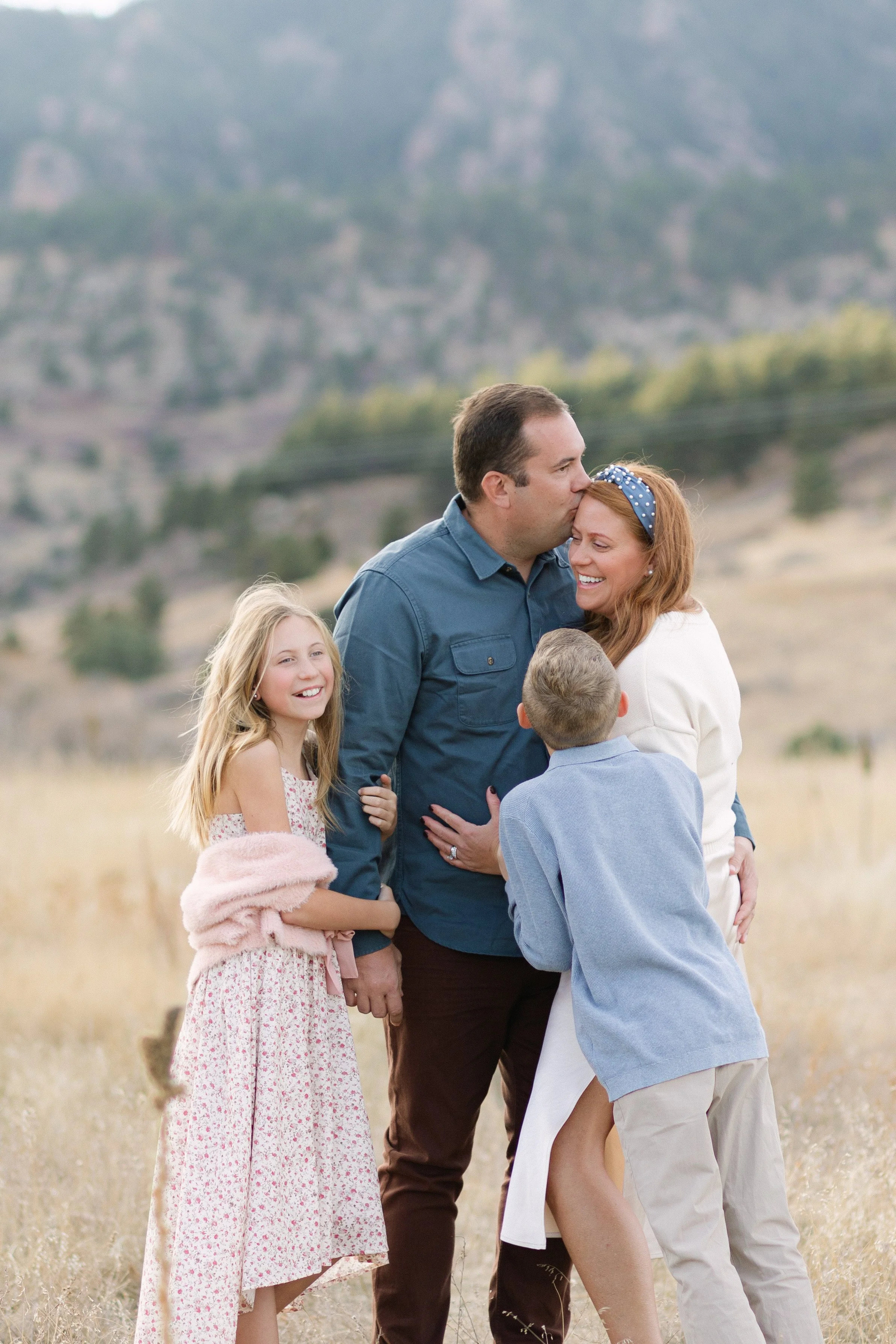 A family of four standing outdoors in a field with mountains in the background. The father is kissing the mother on her forehead, and they are smiling. The daughter and son are also smiling and looking at their parents.
