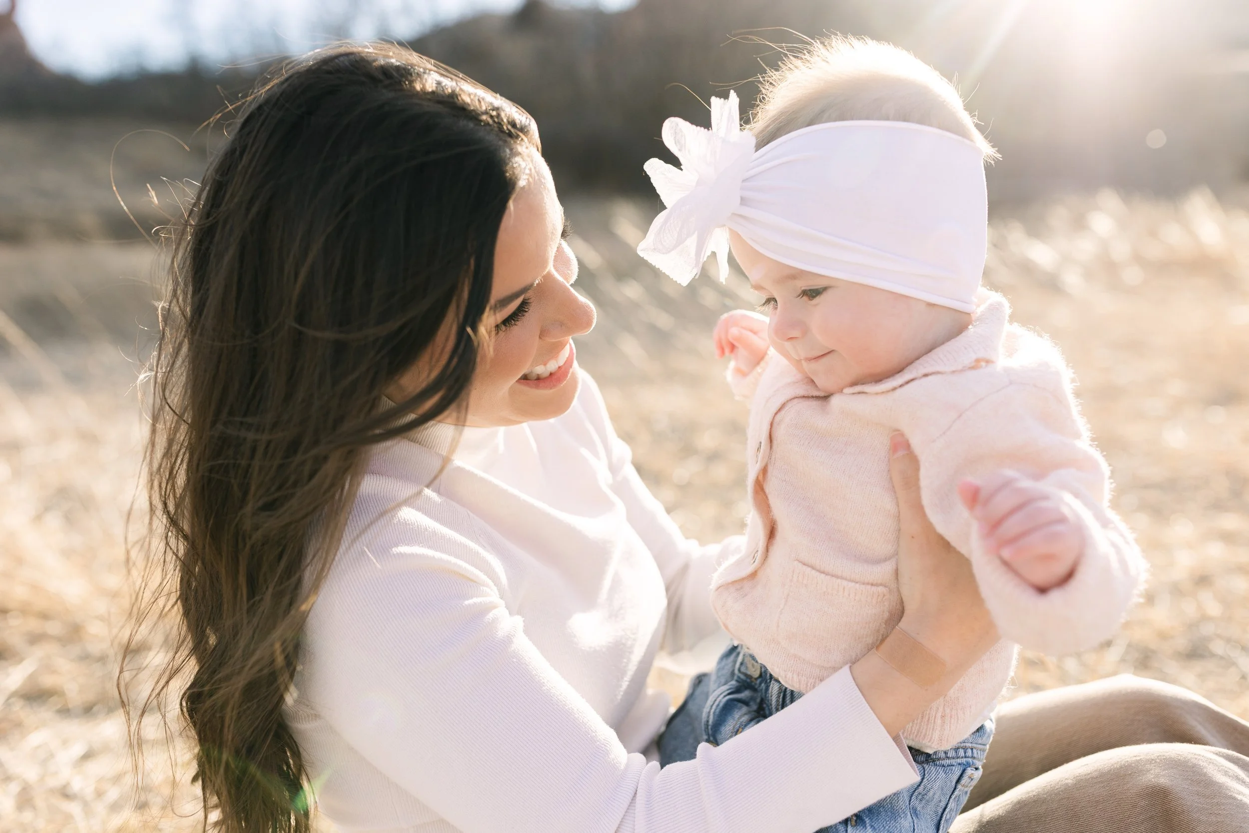 A woman with long dark hair smiling and holding a young girl with blonde hair and a white headband, outdoors on a sunny day.