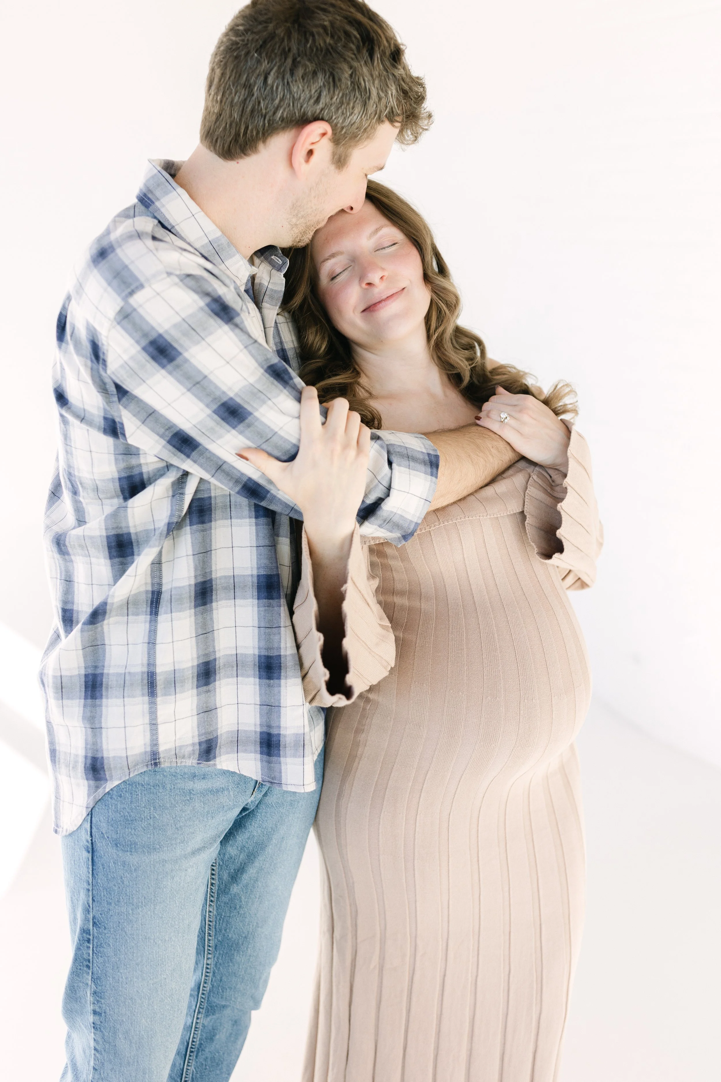 A light and airy maternity photo of an expecting couple embracing in a natural studio setting. The mother rests peacefully in her partner’s arms, highlighting the quiet connection, love, and anticipation of welcoming their baby. This lifestyle matern