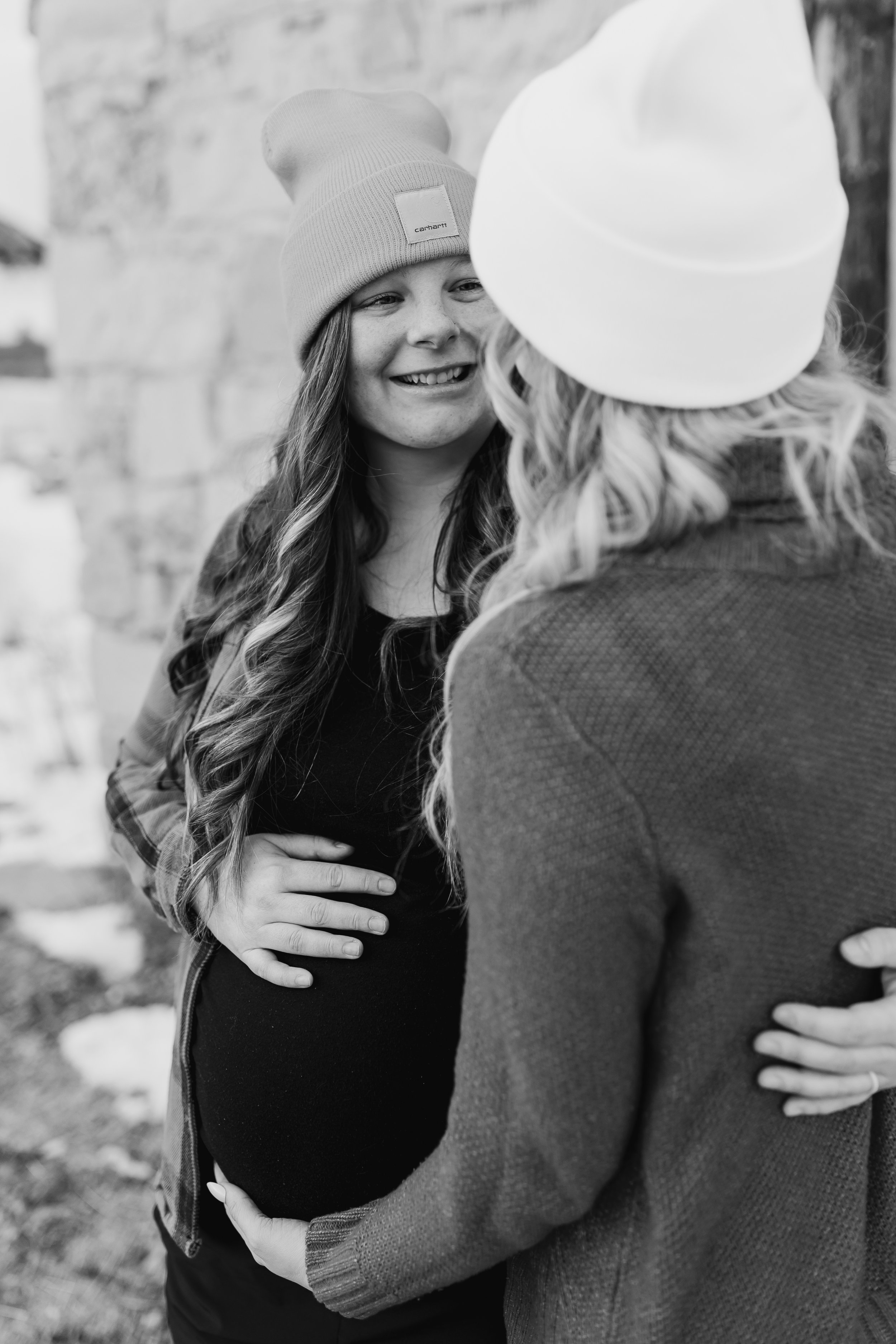 A black and white maternity photo capturing a genuine, emotional moment between an expecting mother and her partner. The mother gently holds her baby bump while sharing a smile, highlighting the connection, anticipation, and love during this special 