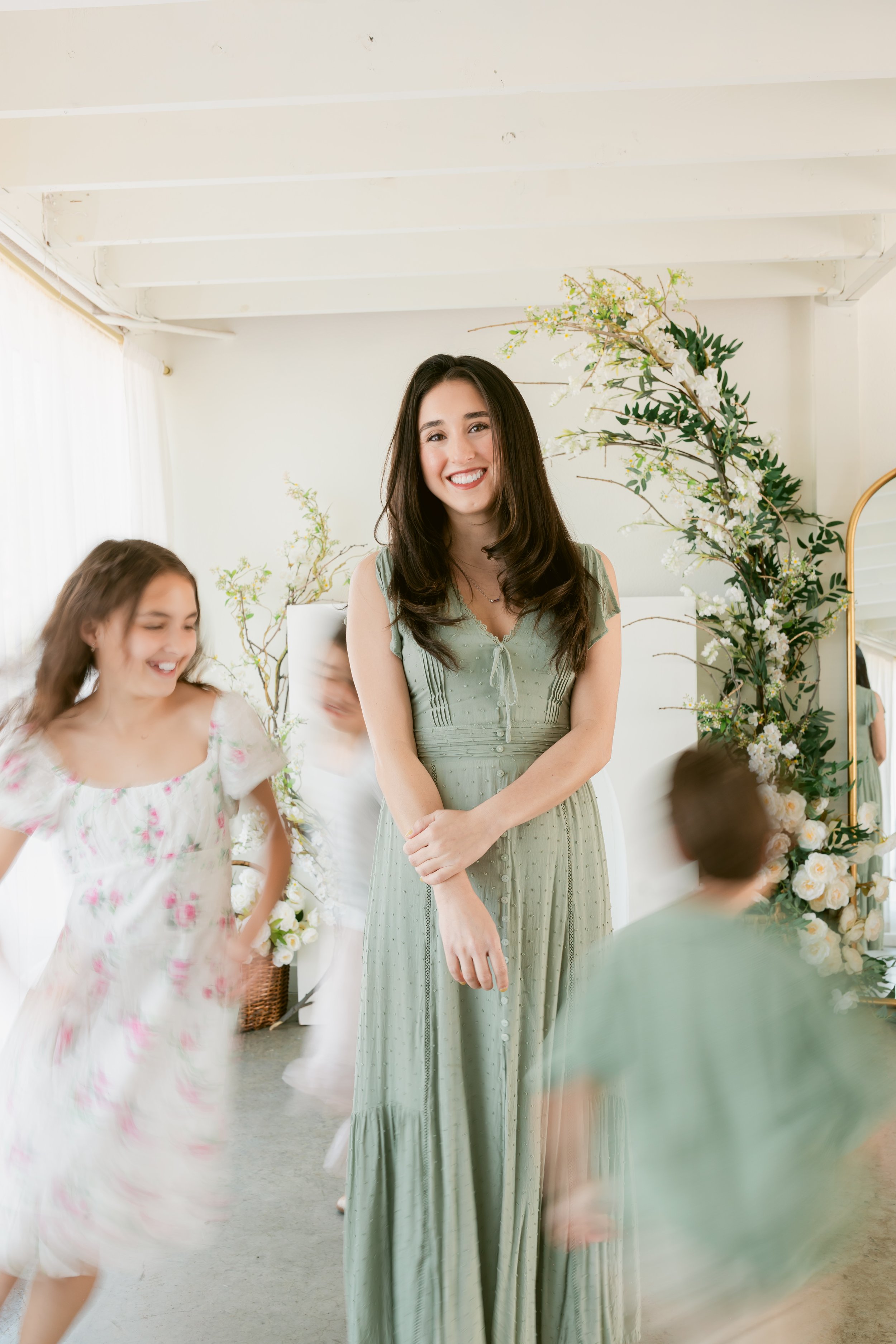 A smiling woman in a green dress standing among children in a bright, floral room.
