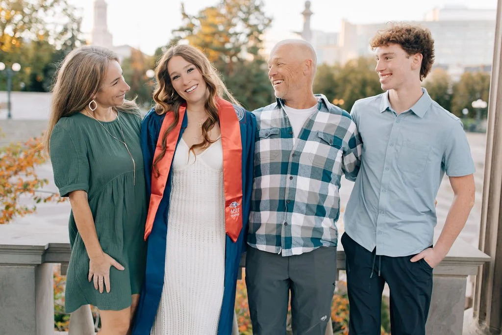 A family standing outdoors, celebrating a graduation. The young woman in the center wears a graduation gown and cap. Her family members are smiling and looking at her, with the background showing trees and buildings.