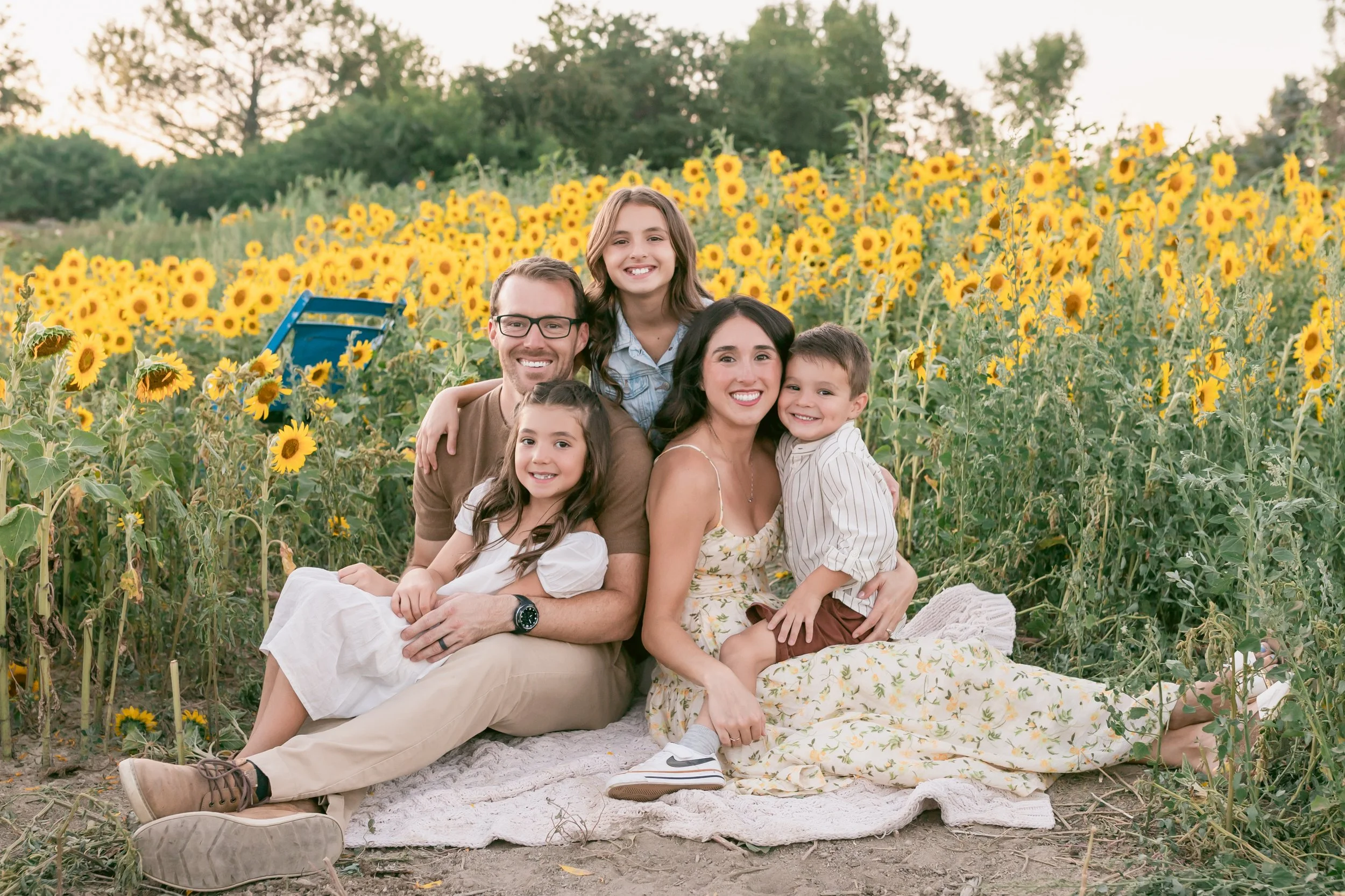Family of five sitting on a blanket in a sunflower field during sunset. The mother and father are smiling, with two daughters and a son, all smiling and embracing each other.
