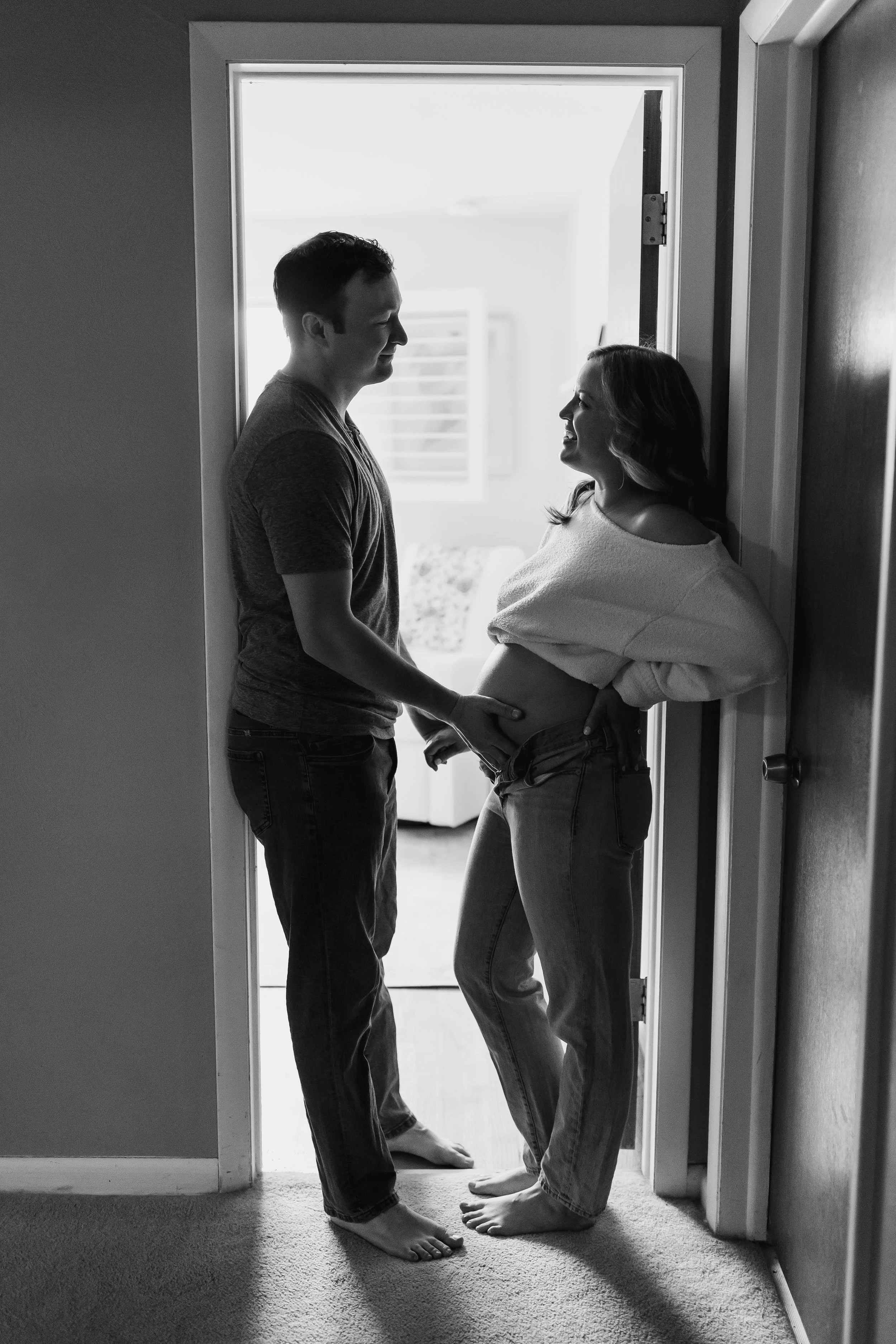 Black and white photo of a couple standing in a doorway, expecting a baby, smiling at each other. The woman has her hand on her pregnant belly.