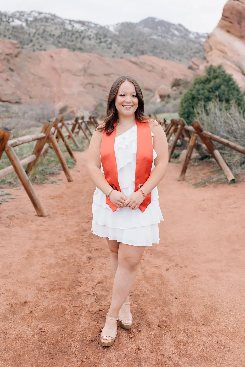 Young woman in white dress with orange stole standing on a dirt path in a desert landscape with red rocks and mountains in the background.
