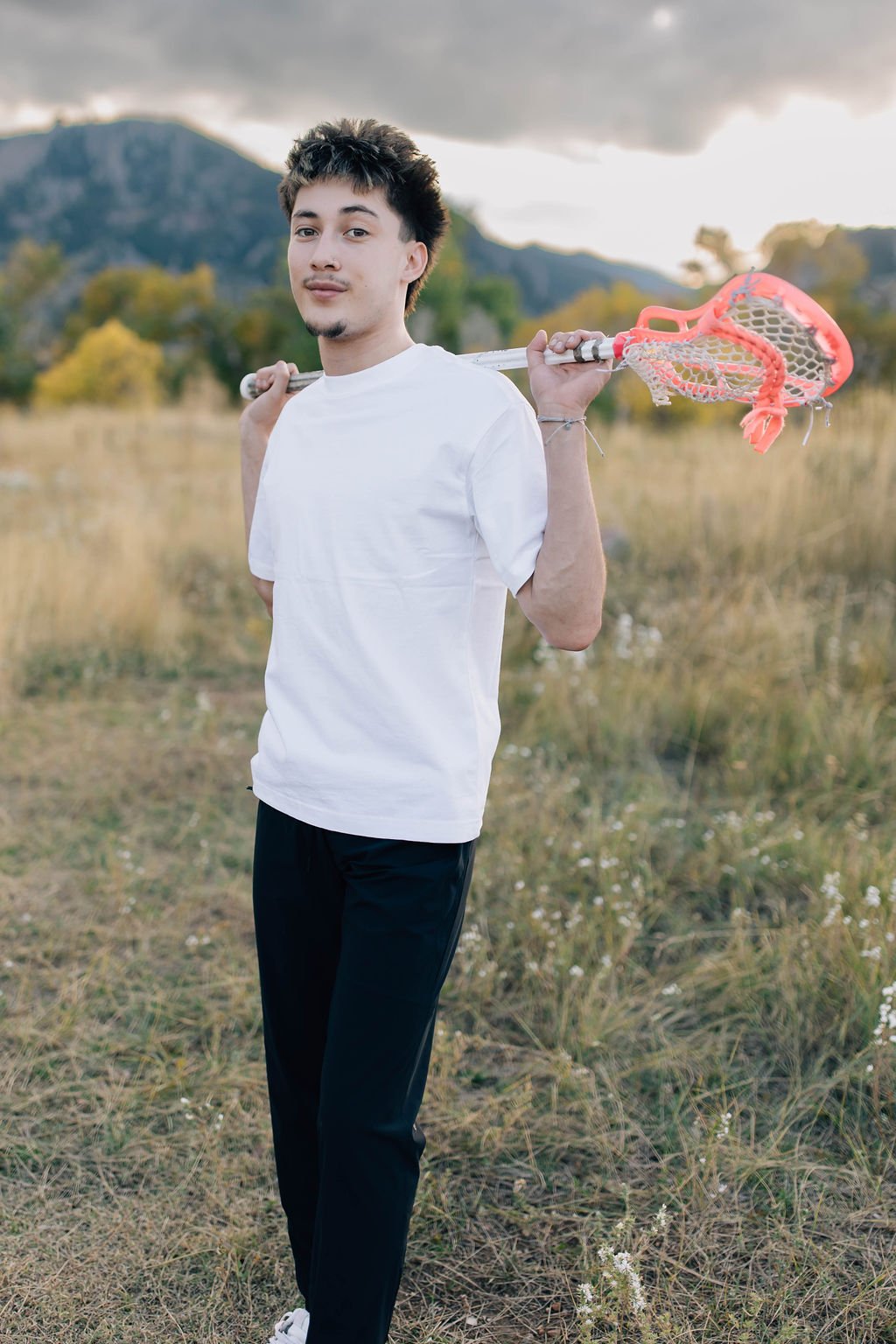 A young man in a white T-shirt and black pants stands outdoors in a grassy field with mountains in the background, holding a mountaineering ice axe over his shoulder.