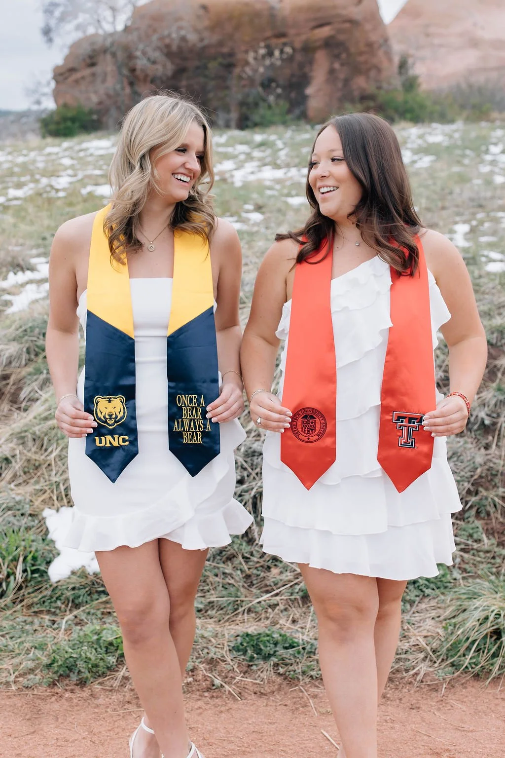 Two women in white dresses wear graduation stoles, smiling at each other outdoors with a rocky landscape and patches of snow behind them.