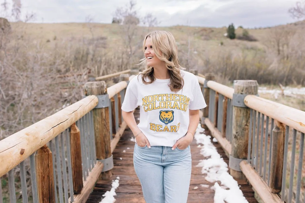 A young woman standing on a wooden bridge with snow on the ground, wearing a white "Northern Colorado Bear" T-shirt and light blue jeans, smiling and looking to her right.