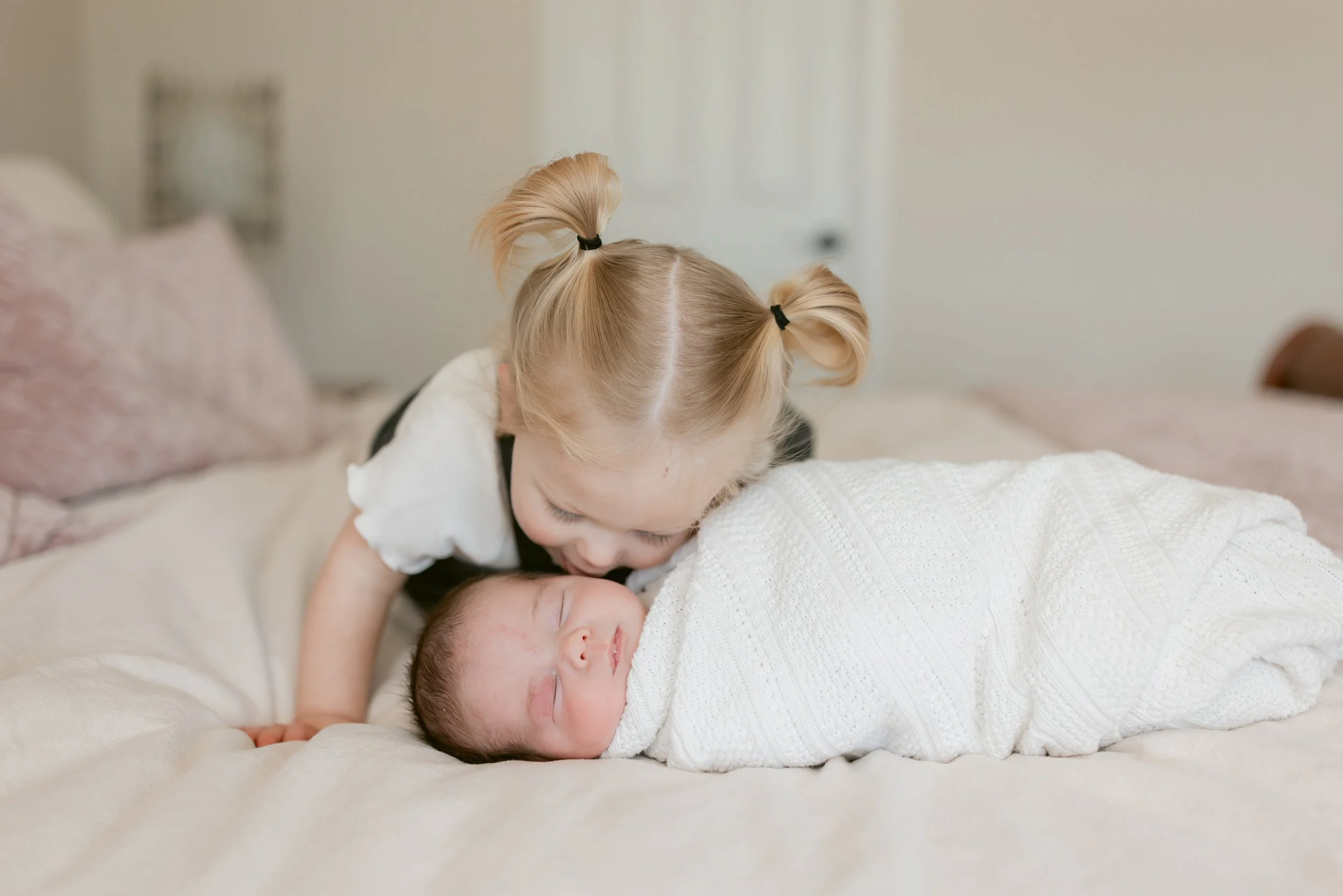 A young girl with blonde hair in two pigtails kisses a sleeping newborn wrapped in white on a bed.