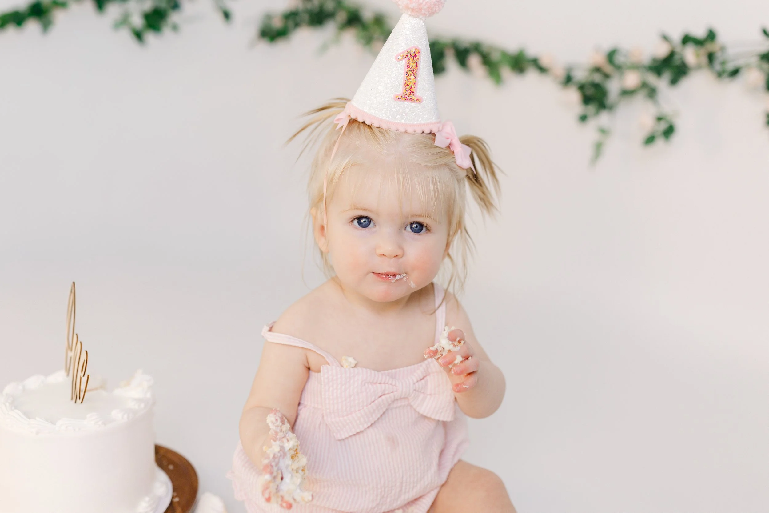A young girl with blonde hair wearing a pink dress and a glittery birthday hat with the number one, eating cake at a birthday celebration.