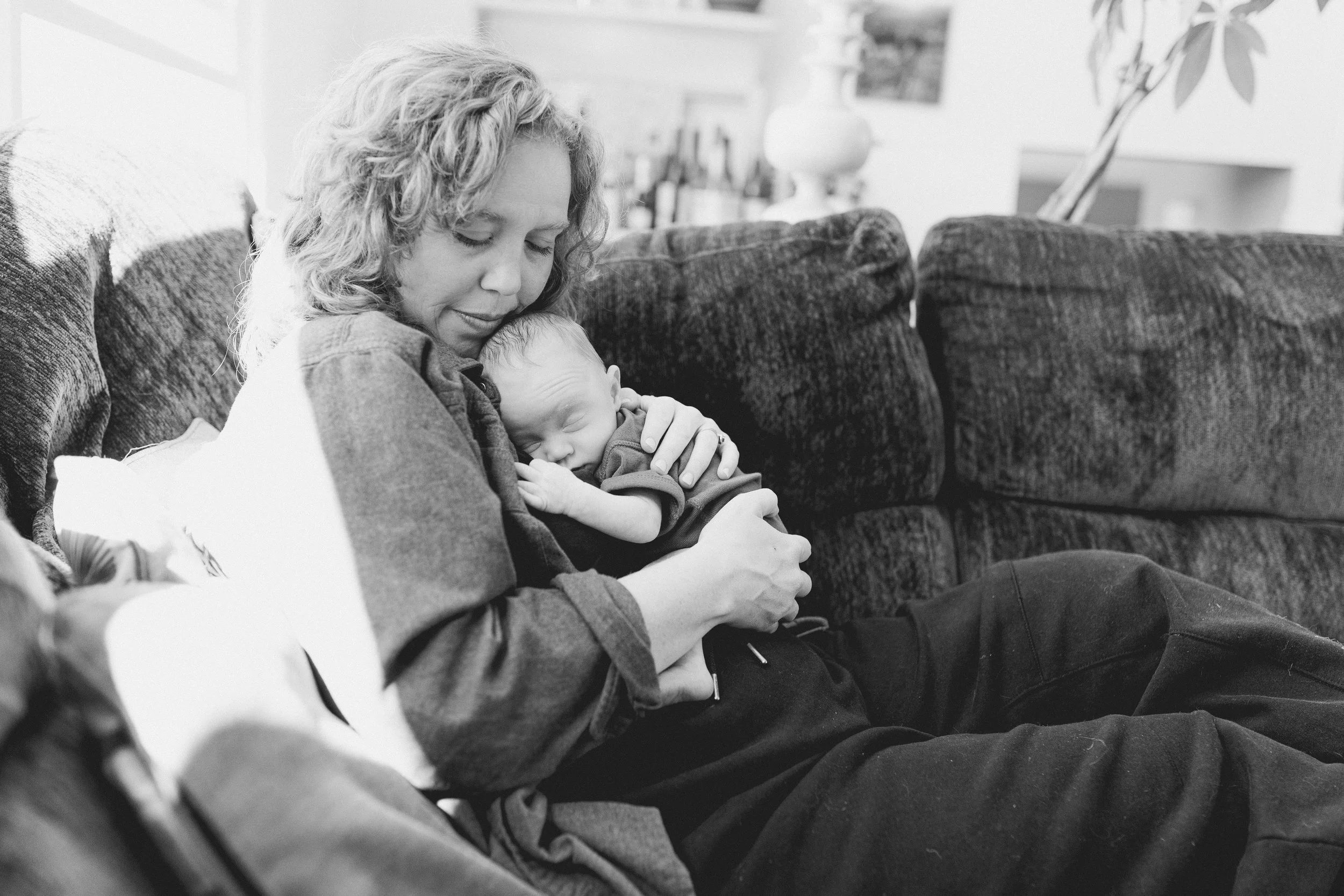 A woman with curly hair sitting on a sofa, holding a sleeping baby close to her chest, both appearing peaceful and relaxed in a cozy indoor setting.