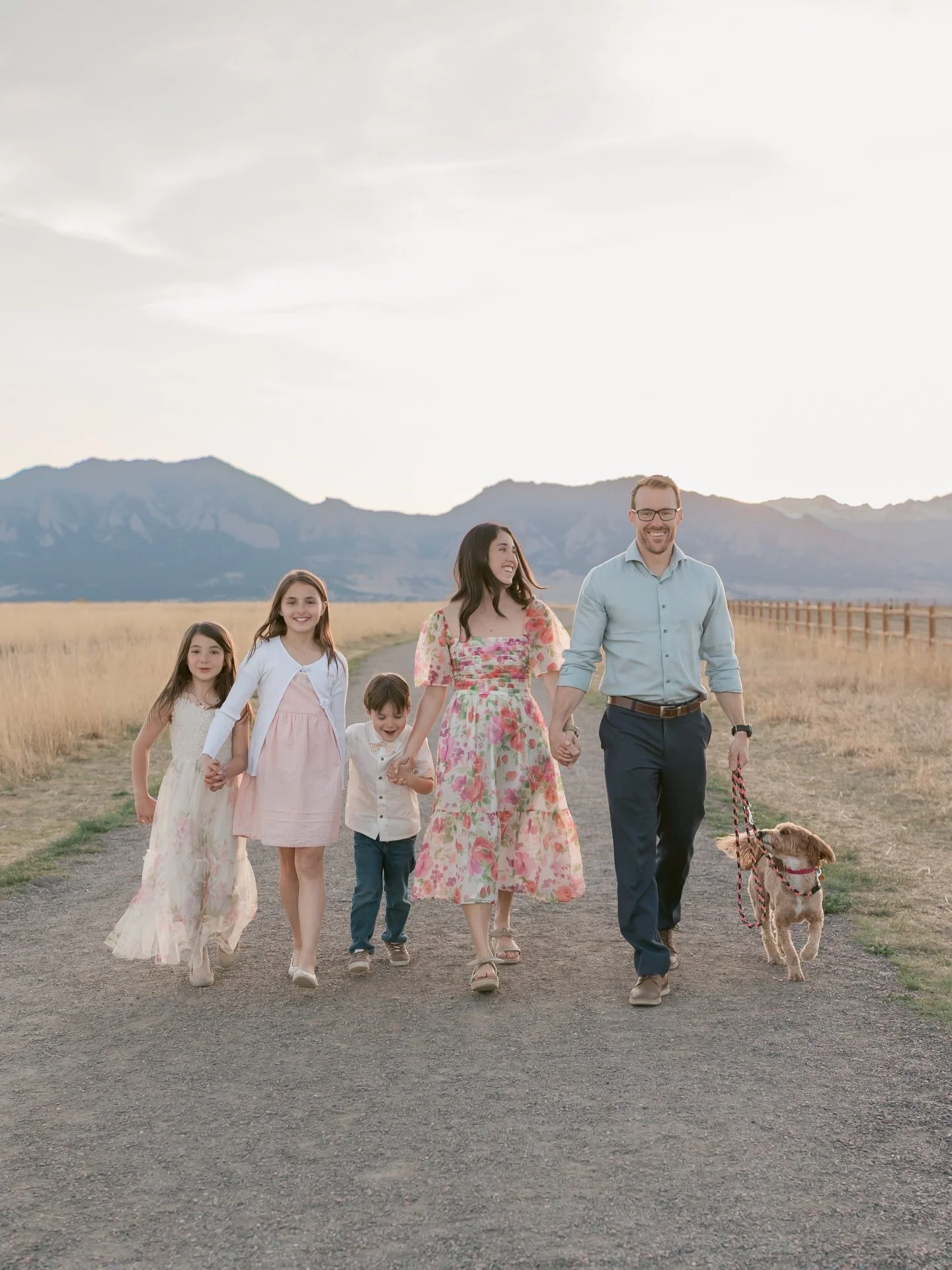 A family of five walking on a gravel path in an open field with mountains in the background, holding hands and smiling, with a dog on a leash.