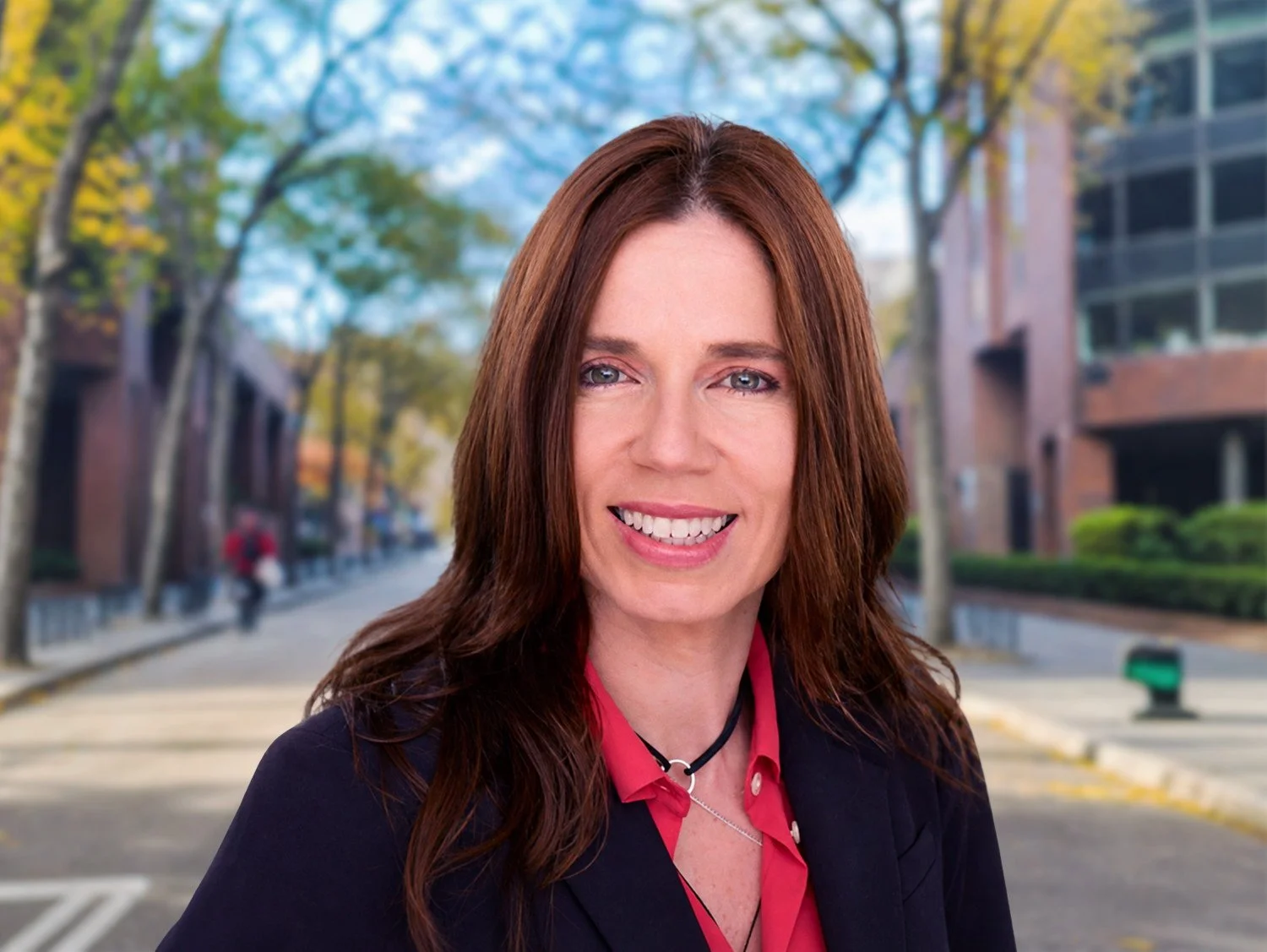 A woman with long red hair and a smile, wearing a black blazer and red blouse, standing outdoors on a city street with trees and buildings in the background.