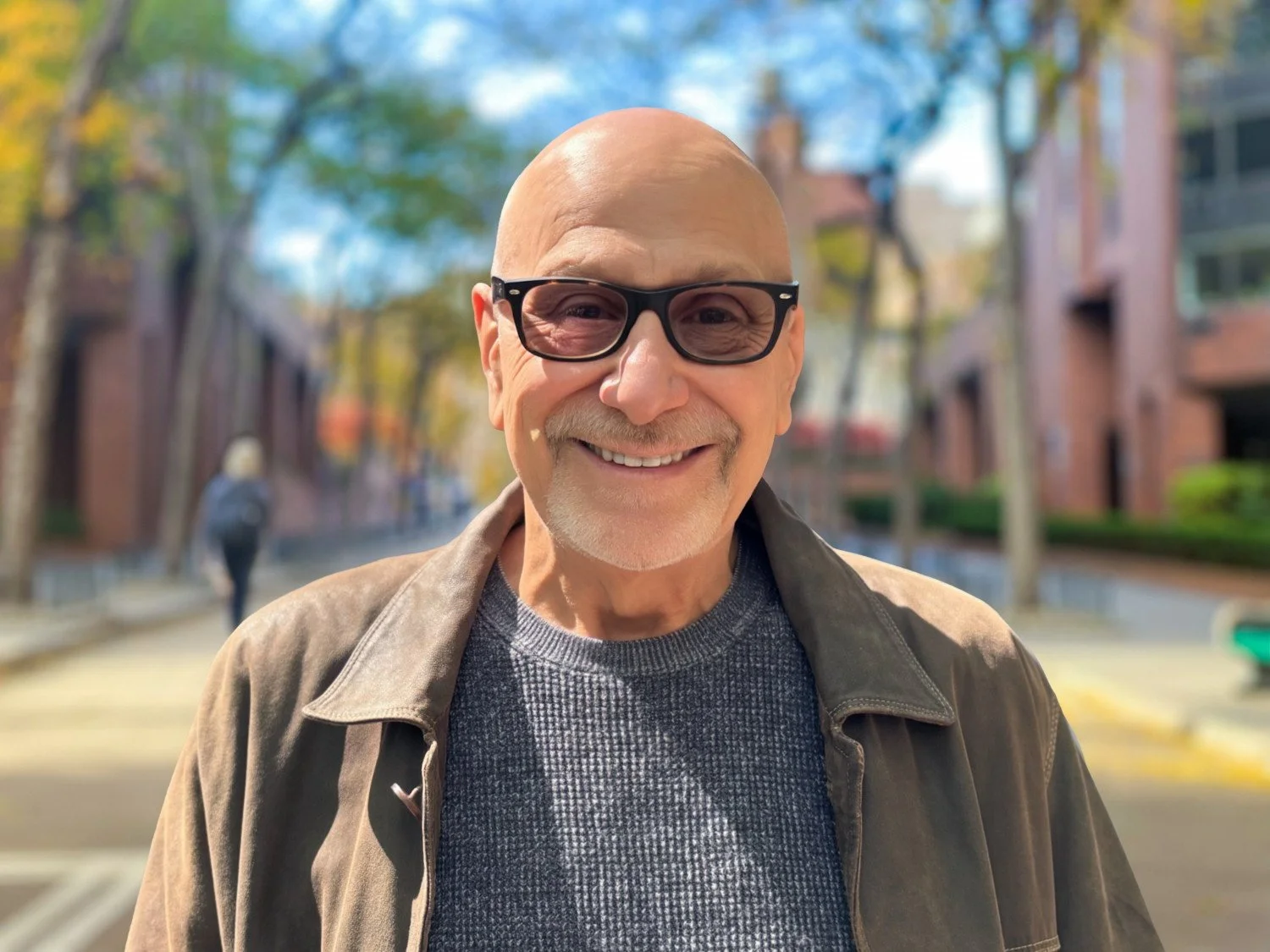 Close-up of smiling older man with bald head, glasses, and casual clothing on a city street with colorful trees and buildings in the background.