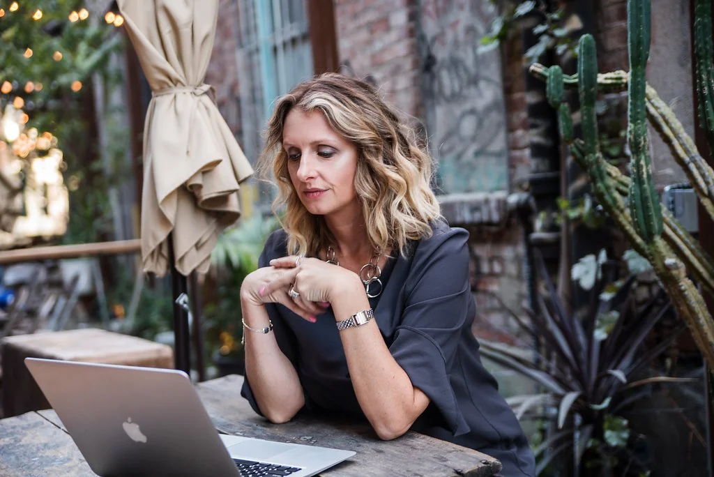 A woman with blonde wavy hair sitting at an outdoor table, looking at her laptop with a pensive expression, surrounded by greenery and brick walls.