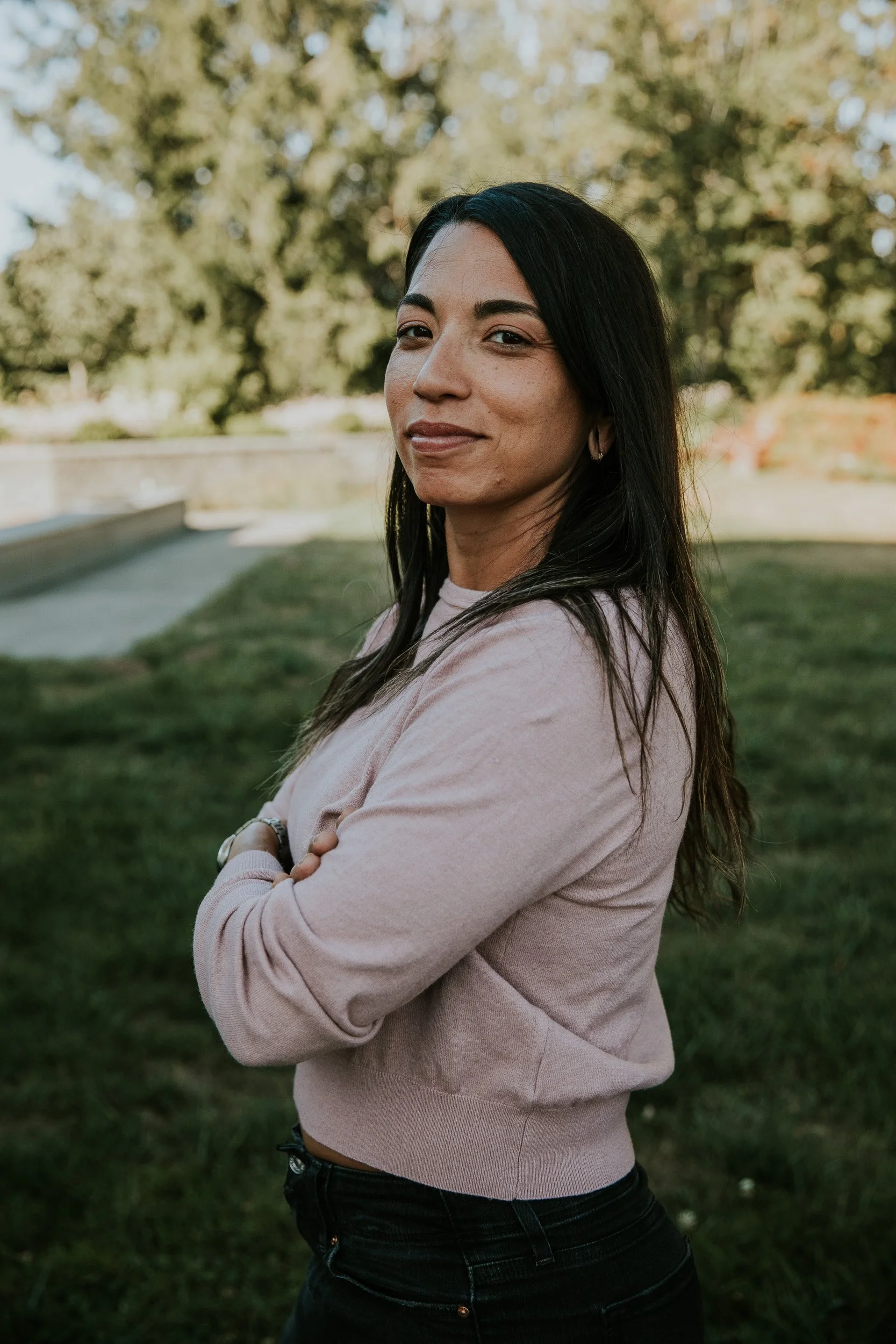 A woman with long dark hair, wearing a light pink sweater and dark jeans, standing outdoors with arms crossed, smiling slightly, with green trees and a stone wall in the background under bright daylight.