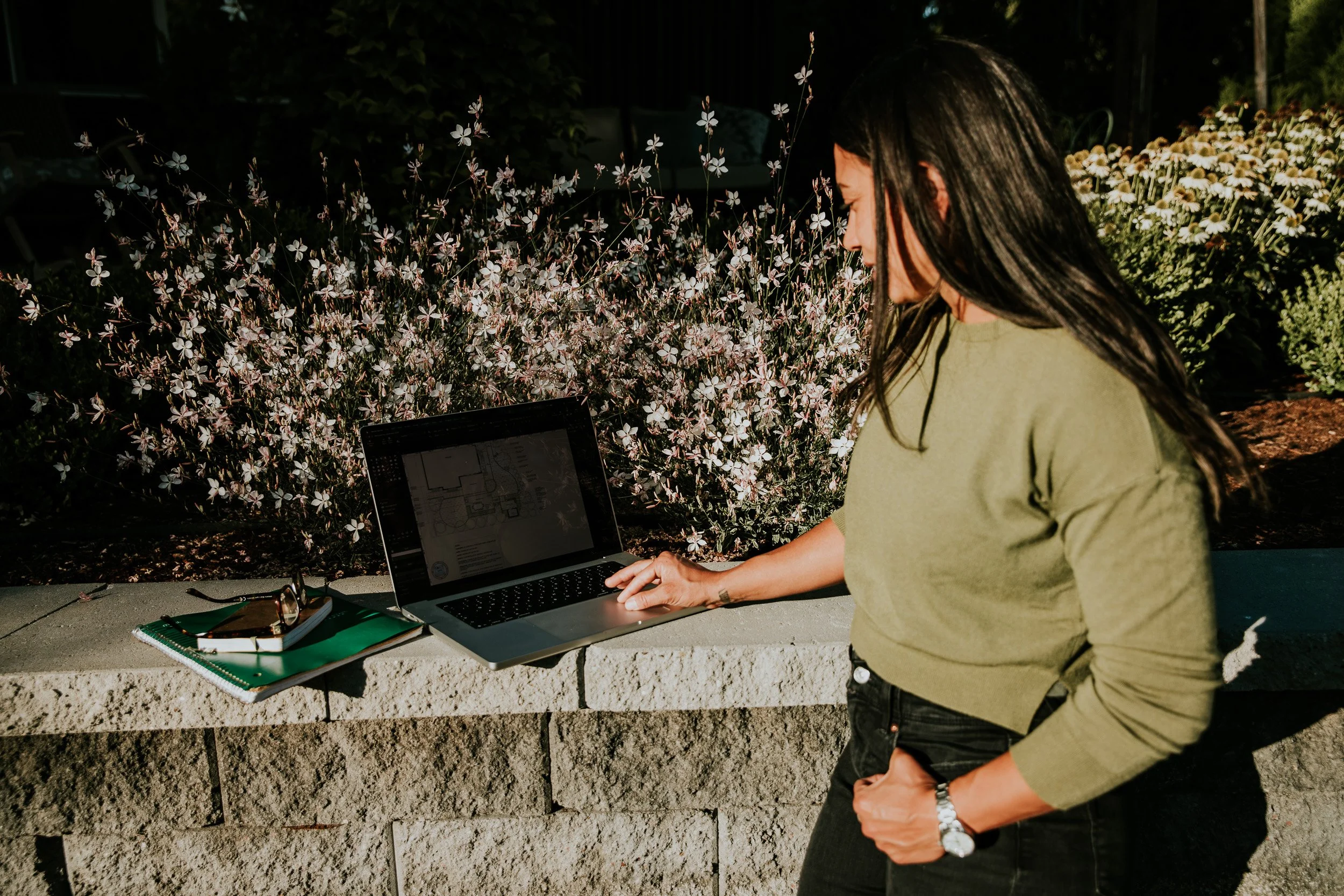 A woman with long dark hair wearing a green shirt and black pants standing outdoors at a table with a laptop, notebook, eyeglasses, and pens, in front of a background of blooming white flowers.