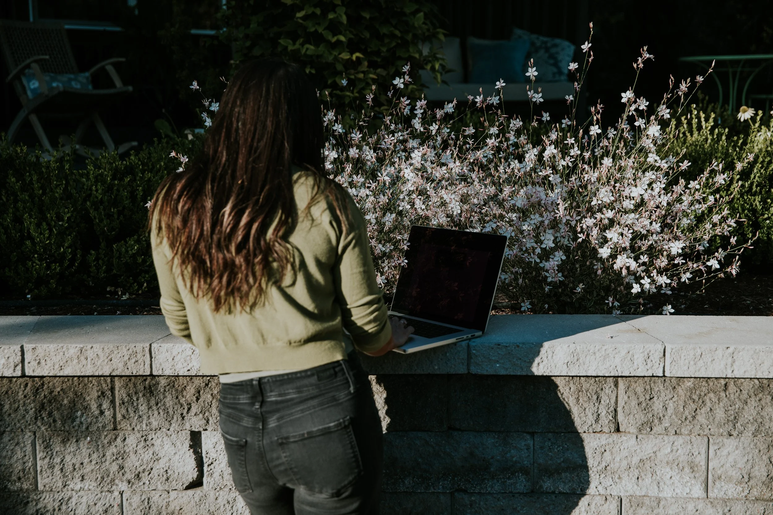 A woman with long dark hair using a laptop outdoors, standing in front of a stone wall with flowering bushes and garden furniture in the background.